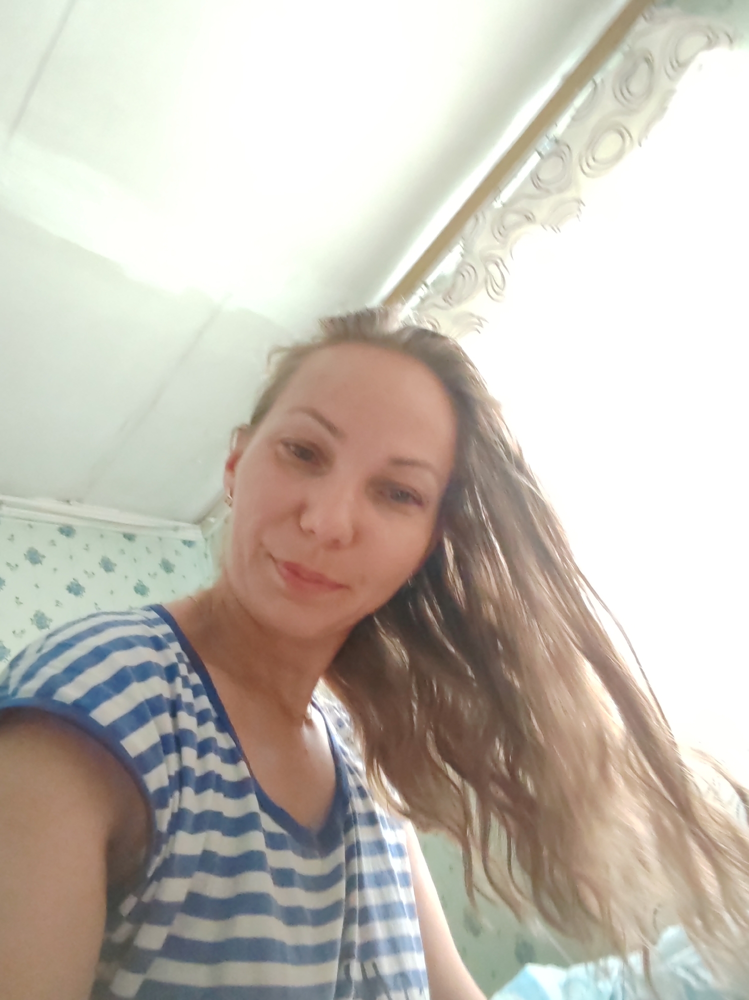 A woman with long, light brown hair is captured in a selfie taken from a low angle. She is wearing a blue and white striped t-shirt. The background appears to be an indoor setting with white ceiling panels and a patterned wallpaper on the wall. The lighting suggests it might be daytime, with a bright light source in the upper right portion of the image. The image does not provide any specific details about the city of Чухлэм, Russia.
