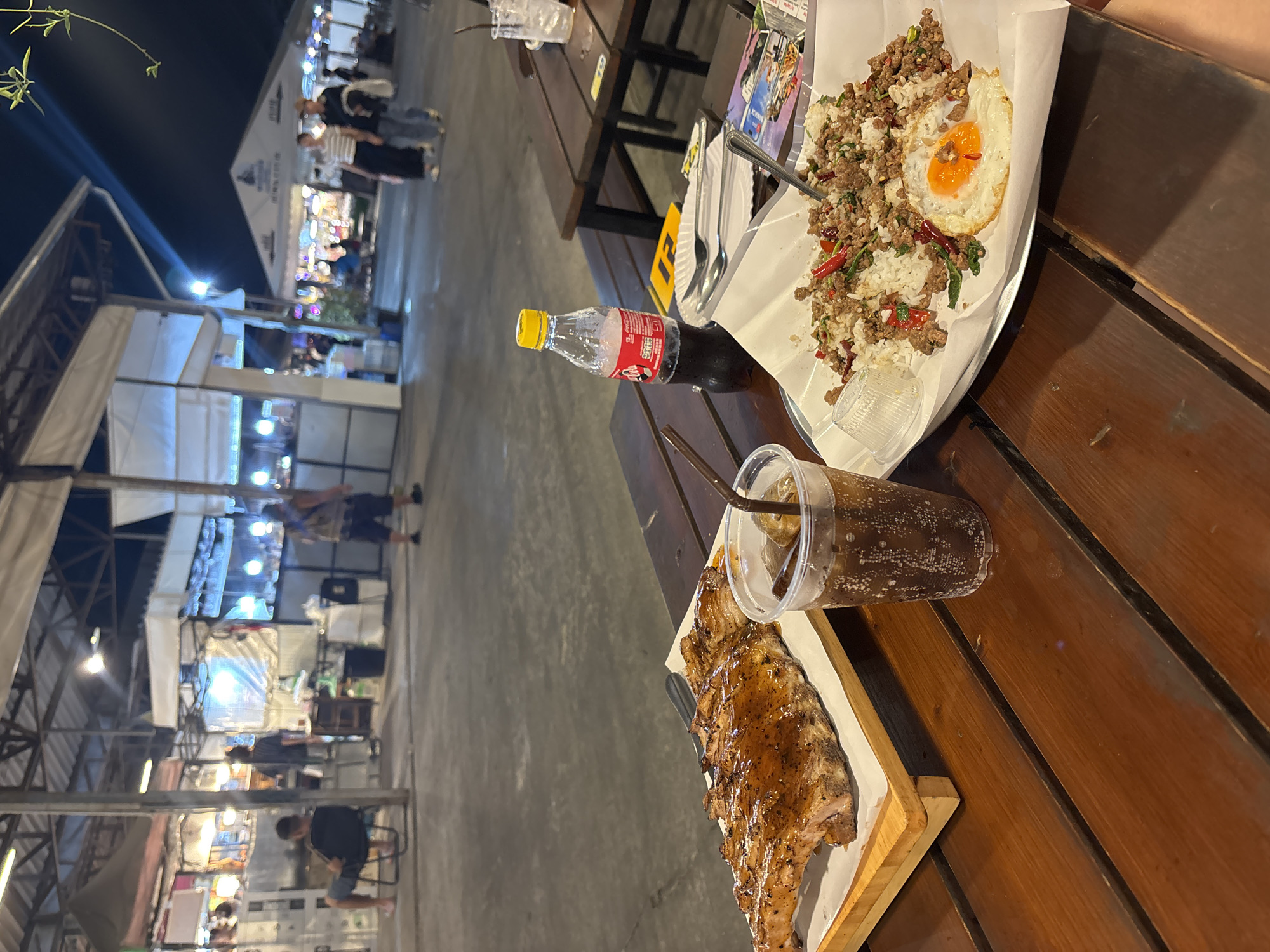 The image captures a vibrant nighttime scene in Bangkok, Thailand, where a person is enjoying a meal at an outdoor food market. A wooden table is laden with dishes and drinks. A serving of Pad Krapow Gai (stir-fried chicken with basil) topped with a fried egg, and a rack of glazed ribs are prominently displayed. A bottle of Coca-Cola and a plastic cup filled with a dark soda complete the meal. In the background, the bustling market comes to life with food stalls illuminated by bright lights, attracting a mix of vendors and customers. The perspective is from the diner's point of view, showcasing the vibrant ambiance of the outdoor market at night.
