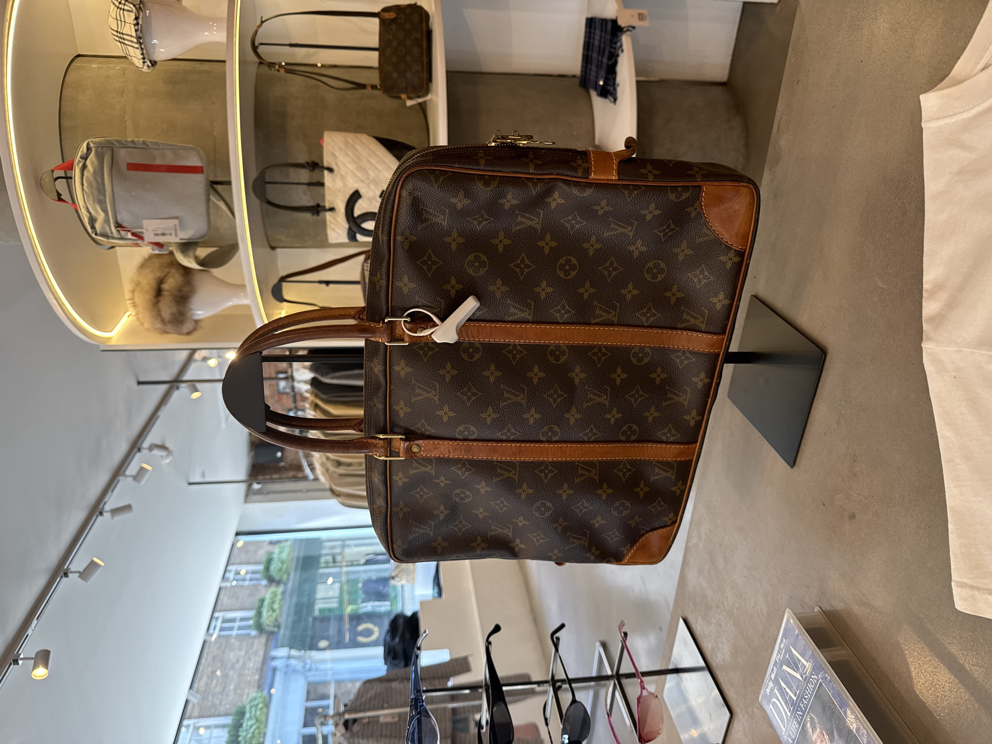 The image displays the interior of a luxury retail store, likely a high-end boutique or consignment shop, featuring a curated selection of fashion accessories. The central focus is a large, brown Louis Vuitton monogrammed travel bag with leather trim and handles, displayed on a black stand. A white anti-theft tag is secured to one of the bag's zippers.
Behind and to the left of the main bag, various other luxury items are showcased on illuminated shelves. These include a black and white checkered cap on a white display head, a grey messenger bag with a red stripe, a fluffy beige hat on another white display head, a smaller brown monogrammed pouch (also Louis Vuitton), and a white quilted bag with a prominent black 'C' logo, suggesting Chanel. Further down, multiple pairs of sunglasses are arranged on a rack.
The store is well-lit with track lighting on the ceiling. Large windows in the background offer a view of an urban outdoor scene, featuring older brick buildings and some greenery, indicating a daytime setting in a city, consistent with London, United Kingdom. On a surface to the right of the main bag, a plain white t-shirt is visible, alongside a book titled "DIANA A LIFE IN FASHION." The scene depicts a static retail display of designer goods.