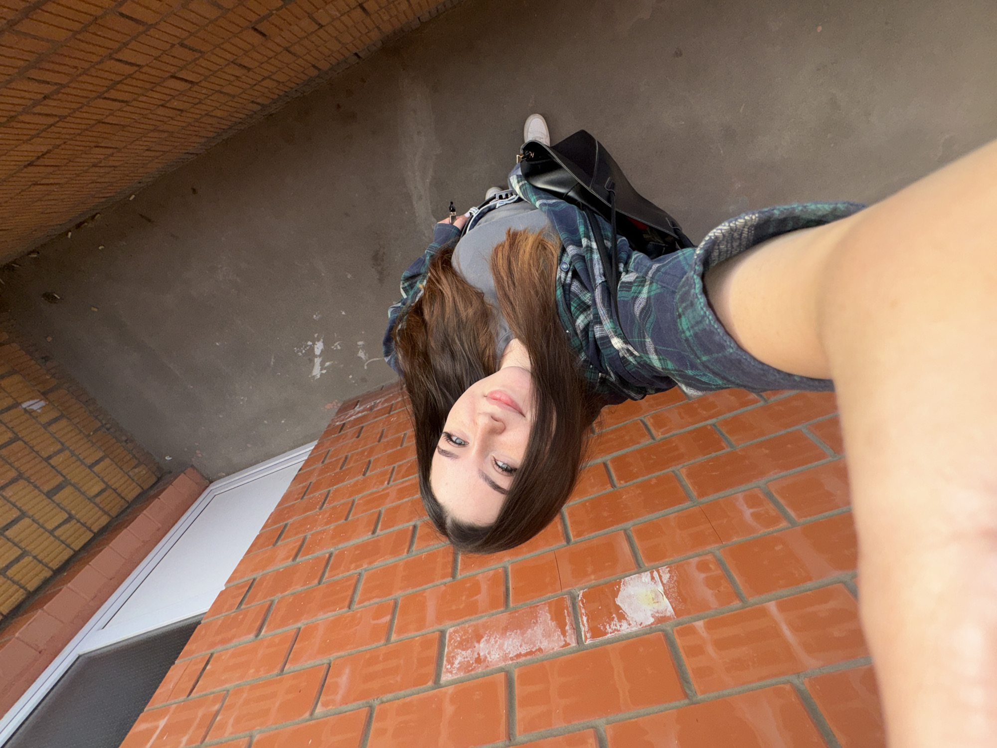 A young woman is taking a selfie outdoors, standing next to a brick wall. She has long, dark brown hair and is wearing a green and blue plaid shirt over a grey t-shirt and grey pants. She is holding a black bag. The background consists of a grey concrete ground and a brick wall. The image is oriented vertically and appears to be taken during the daytime. There is no information in the image to suggest the location is the city of Perm, Russia.