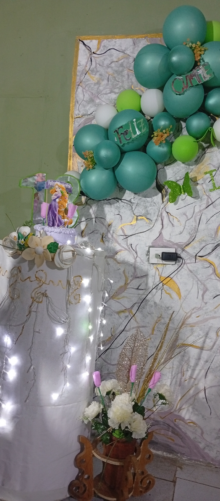 A birthday celebration is captured indoors. A backdrop with swirling gray and gold designs is partially covered with clusters of teal and white balloons, some adorned with floral accents and "Feliz" and "Cumple" cutouts. A table draped in white, adorned with fairy lights and a cake featuring Rapunzel, adds to the festive atmosphere. A floral arrangement sits on an ornate wooden stand. A wall outlet with a plugged-in black adapter is visible in the background.