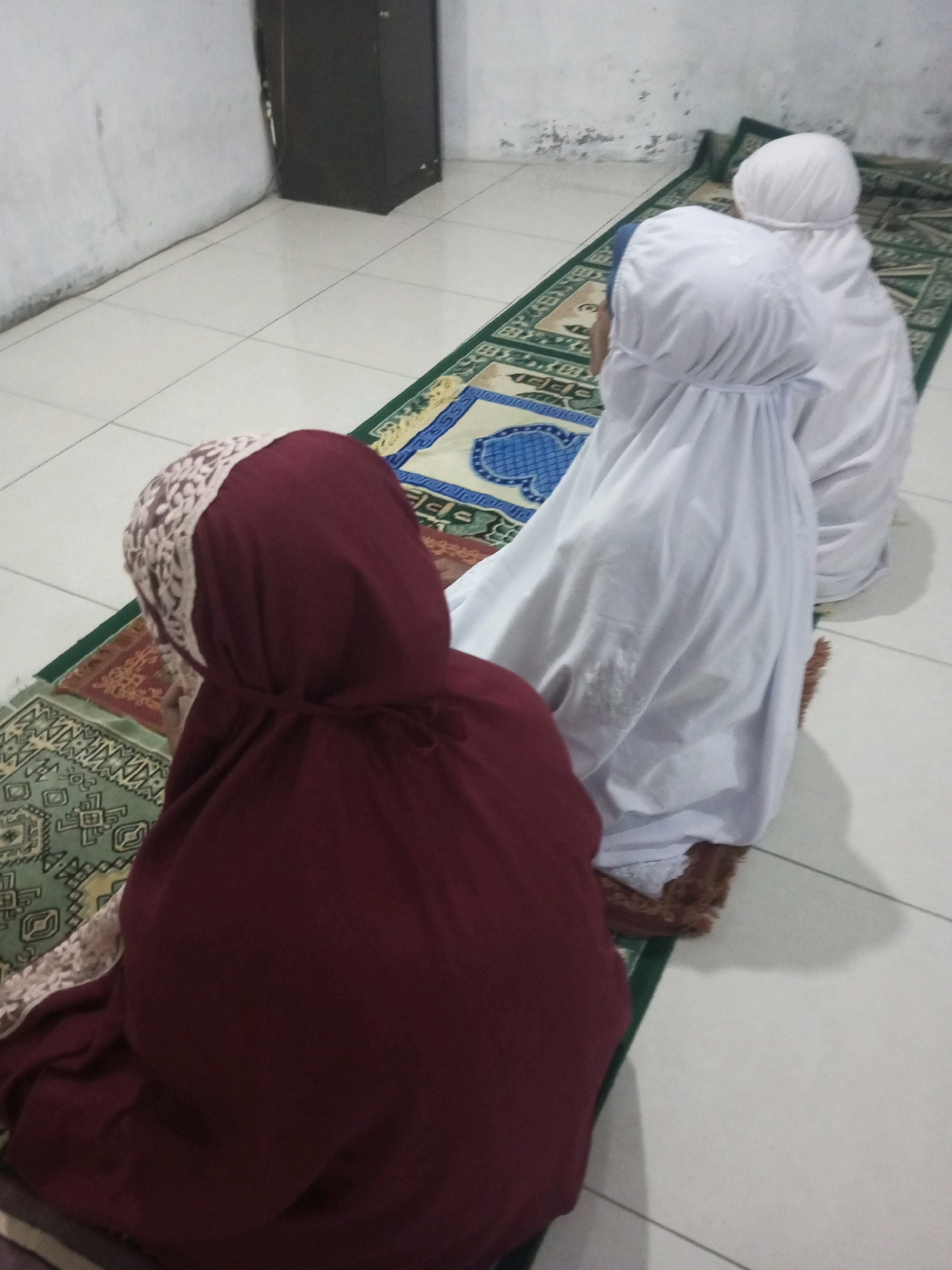 Three individuals, appearing to be children, are engaged in prayer on prayer mats on the floor of a room. The first child in the foreground wears a maroon hijab and a matching garment. The second child, positioned behind the first, wears a white hijab and prayer garment. The third child, in the background, also wears a white hijab and prayer garment. They are facing away from the camera and towards the right of the image. The floor is tiled in a light color. A dark, tall cabinet is visible in the background to the left. The prayer mats have intricate patterns in various colors, including green, blue, red, and yellow. The scene takes place indoors, likely in a residential setting in Andongsili, Indonesia.