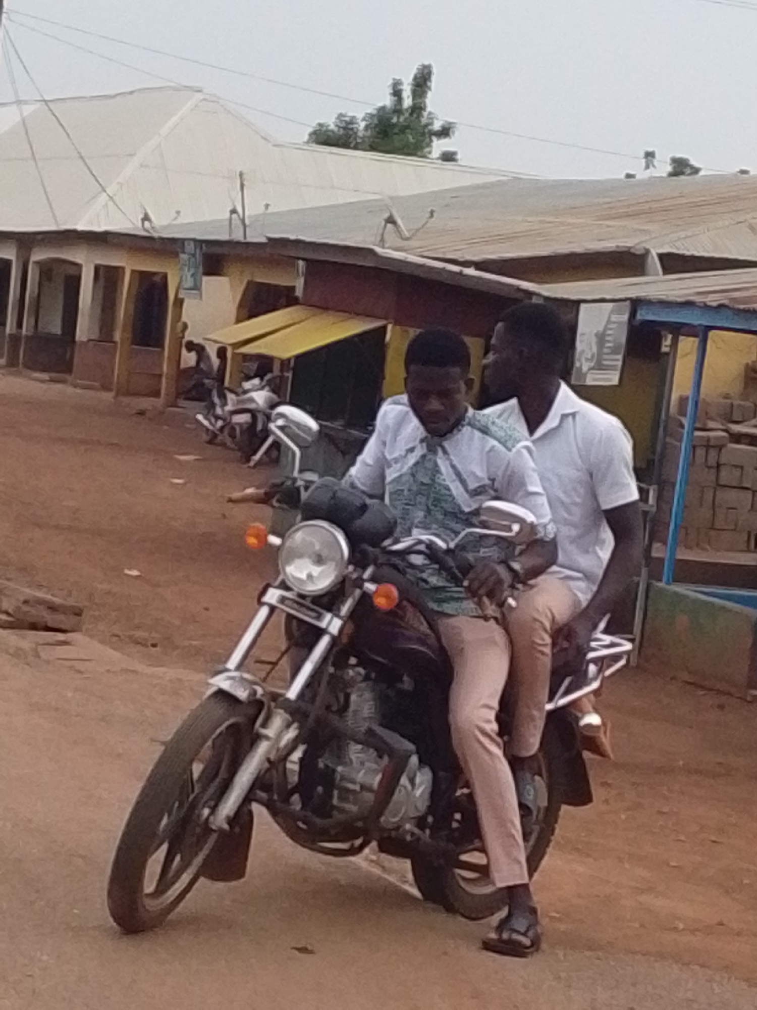 Two African men are riding a motorcycle. The driver is wearing a patterned shirt and light-colored pants, and the passenger is wearing a white shirt and light-colored pants. Both are wearing sandals. The motorcycle is in the foreground, and a street scene is in the background with several buildings, including one with a yellow awning. There are bricks on the right of the shot. The image appears to be taken during the day. Based on the style of buildings and general appearance, the location is likely Tamale, Ghana.