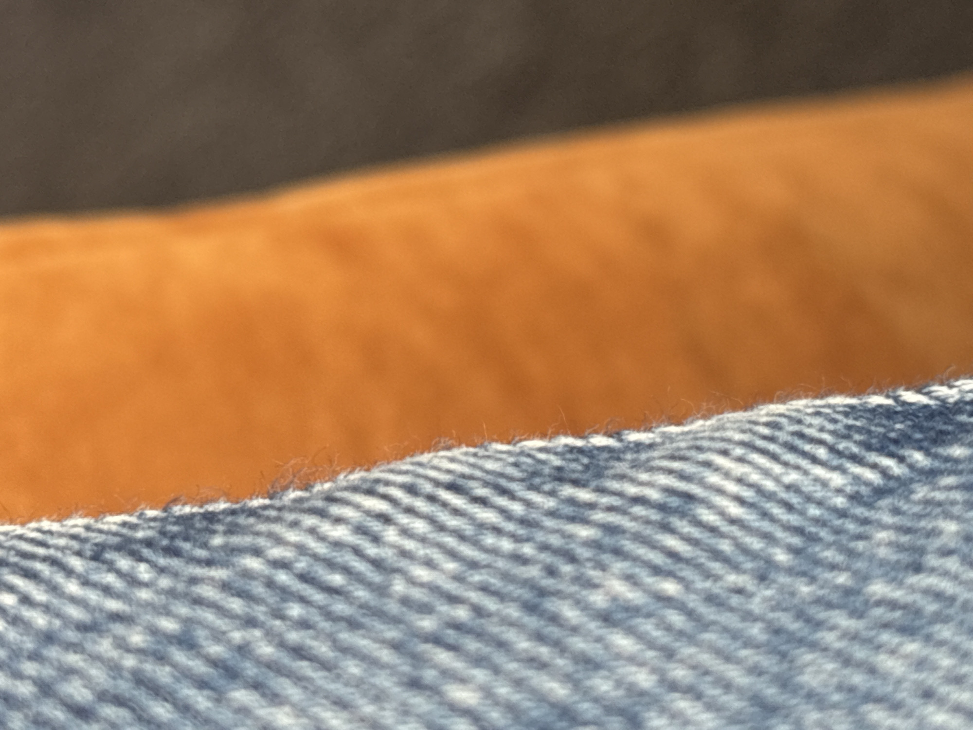 The image is a close-up, abstract shot of fabric textures. On the left, a blue denim fabric with a visible woven pattern is sharply in focus. To the right of the denim, a soft, blurred orange fabric occupies the middle of the frame. Further to the right, a dark, blurred background, possibly another surface or shadow, is visible. The focus is on the juxtaposition of the sharp denim texture against the soft, out-of-focus orange and dark areas. There are no people or identifiable landmarks present. The image is entirely composed of fabric textures.