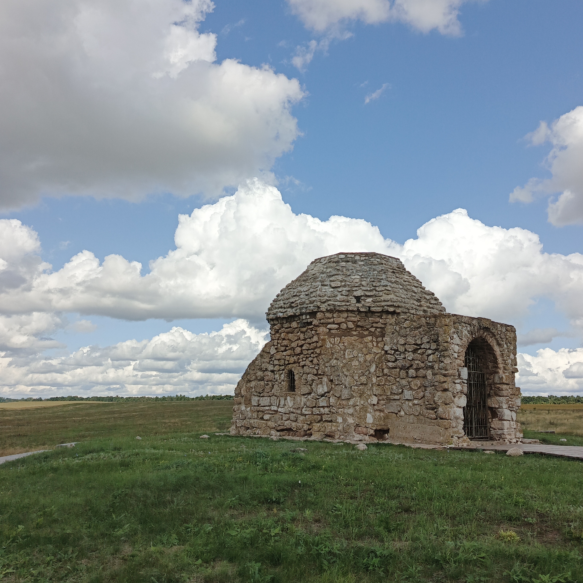 An ancient stone structure, possibly a tomb or mausoleum, stands on a grassy hill under a partly cloudy blue sky in Verkhnie Termy, Russia. The structure is round with a domed roof made of rough-hewn stones. The walls are constructed from similar stones, with a small arched entryway covered by a metal grate on the right side and a small, barred window on the left. The surrounding environment is a green, gently sloping field. In the background, a flat, distant landscape with trees and fields stretches to the horizon under a dramatic sky filled with large, white, and grey cumulus clouds.