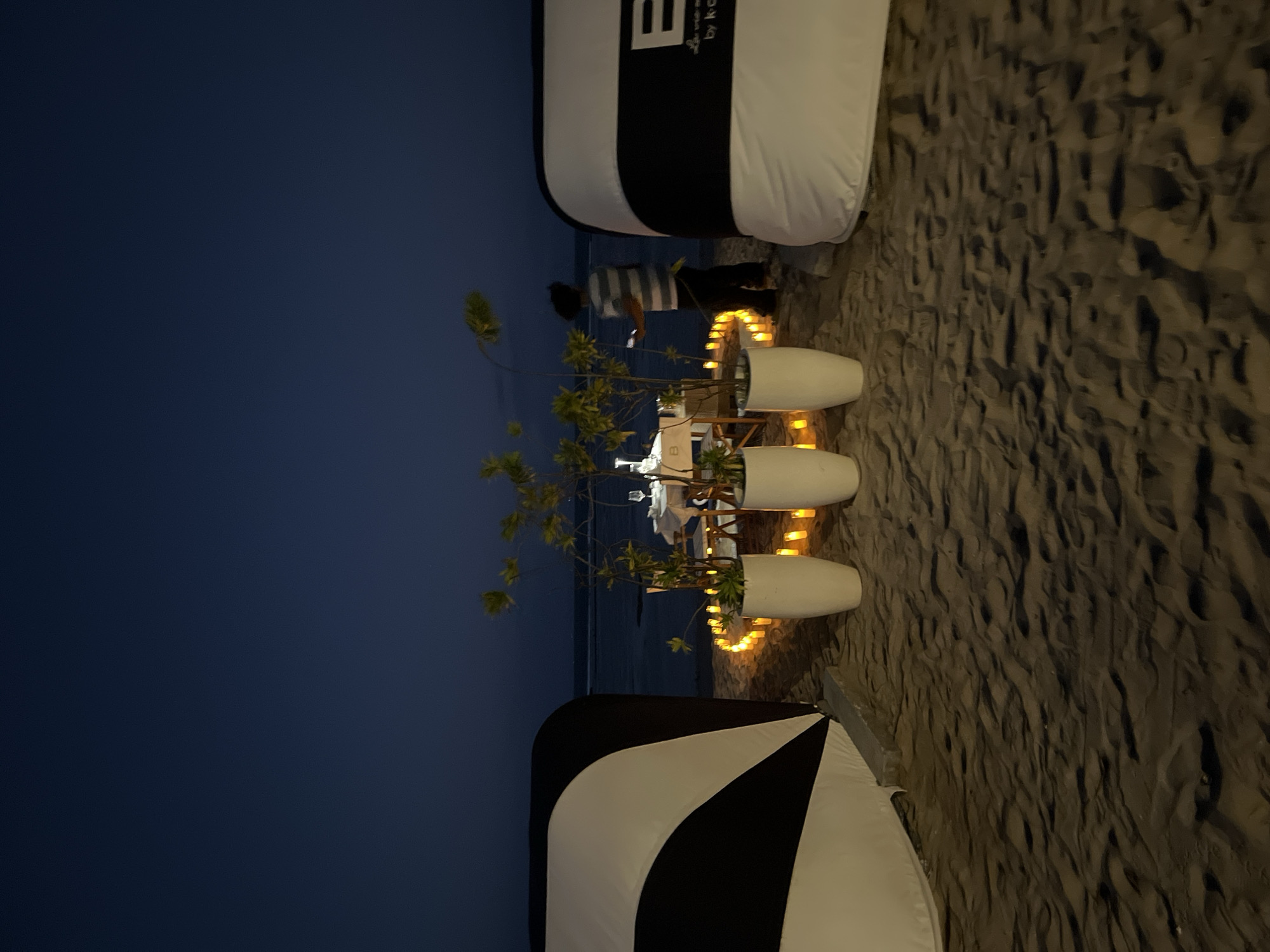 At dusk on a sandy beach in Kuta Selatan, Indonesia, a romantic dinner setting is arranged. Two large, black and white striped structures, resembling tents or marquees, are positioned on the left and right sides of the frame, framing the scene. In the center, a round dining table is set with white tablecloths, chairs, and wine glasses, illuminated by a circle of numerous glowing candles on the sand. Two large white planters, each containing a small, leafy tree, stand in front of the table. A person wearing a striped t-shirt and dark pants is visible in the background, to the right of the table, appearing to be attending to the setup. The ocean is visible in the distance, reflecting the twilight sky. The sand has a textured, rippled surface, indicative of a beach environment.