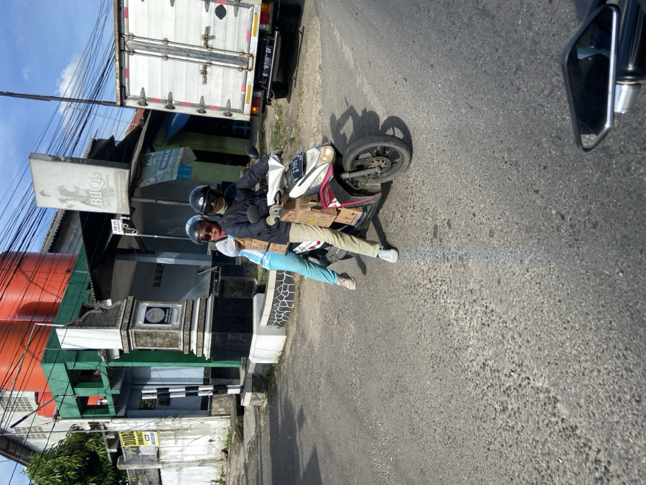 A man and a woman are riding a motorbike on a paved road in Jatiluhur, Indonesia. The man is driving and the woman is sitting behind him. The motorbike has several cardboard boxes stacked on the back. The road is empty. In the background, there are some buildings, including a white truck with a license plate "E 8106 BF". The sky is blue and the sun is shining. The time is daytime. The weather is sunny and clear. The motorbike is a Honda Vario.