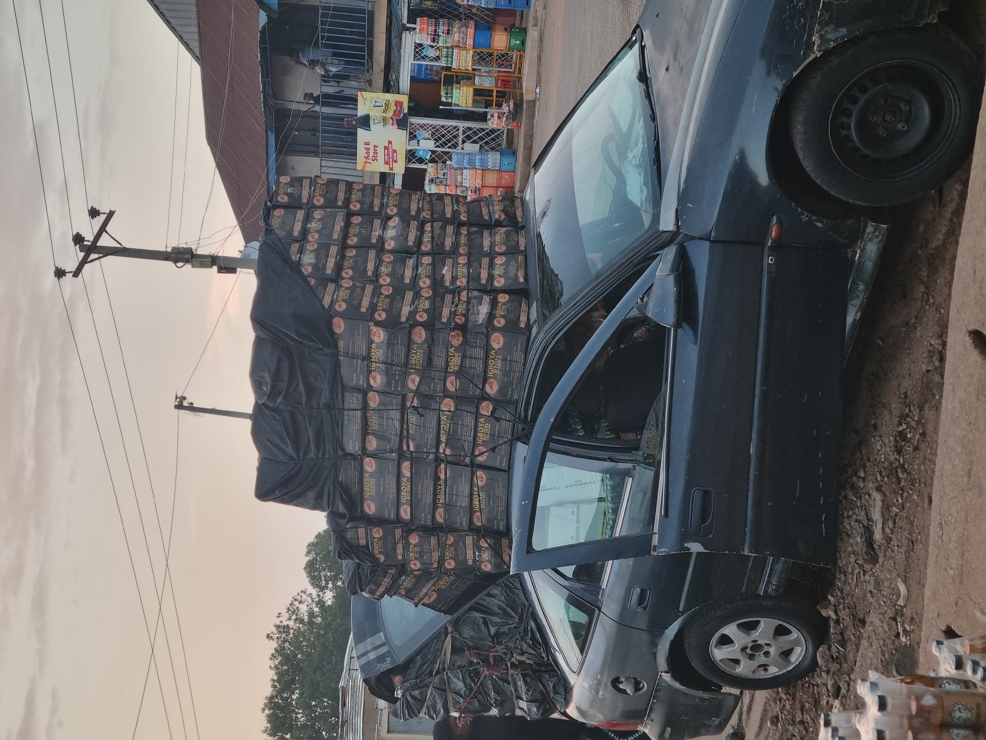 Here is a description of the image:
A black sedan is parked on a rough-hewn curb with its door open, heavily overloaded with stacks of packaged goods labeled "IGBOYA BITTERS". The vehicle is adjacent to a small storefront identified as "And B Store" located in Jos, Nigeria. The store displays various beverages. Overhead power lines are visible against a muted sky.