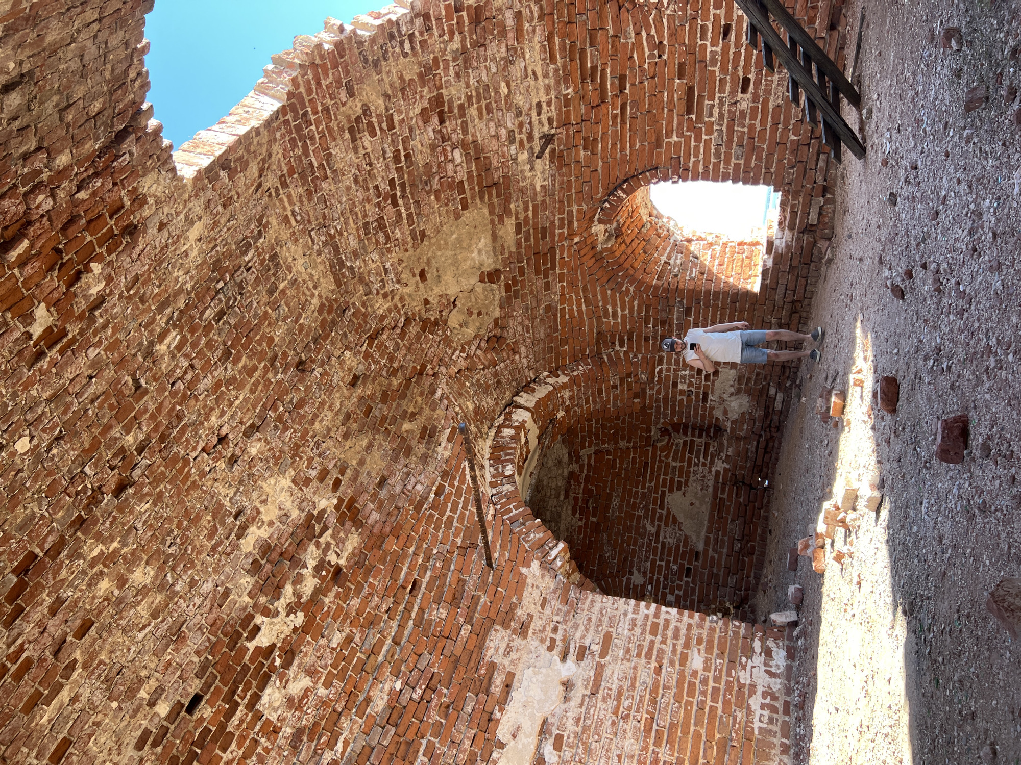 The image depicts the interior of a brick structure, likely a ruin, in Шуран, Russia. The walls are constructed of red brick and show signs of age and deterioration, with missing sections and crumbling mortar. Several arched openings are visible in the walls. A person is standing within the structure, near one of the openings. A partial view of wooden steps is present. The ground is dirt and debris. The sky is visible through openings in the structure.