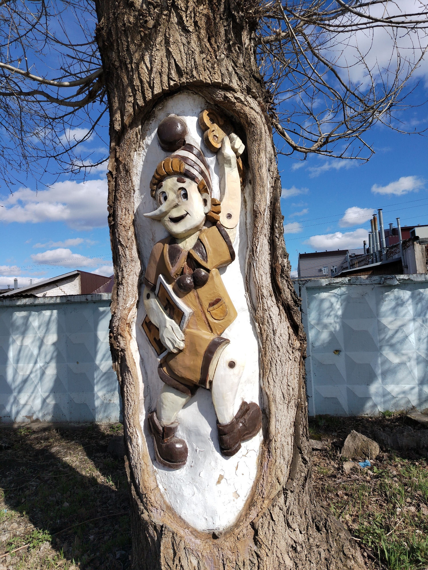 A carving of Pinocchio has been embedded into a tree trunk. He is wearing a striped hat and a brown outfit, and holding a key and a book. The tree is situated in Chaikovsky, Russia, near a light blue wall and some buildings with chimneys. The sky above is blue with scattered clouds, and bare branches extend from the tree. The ground around the tree is covered with grass and debris.