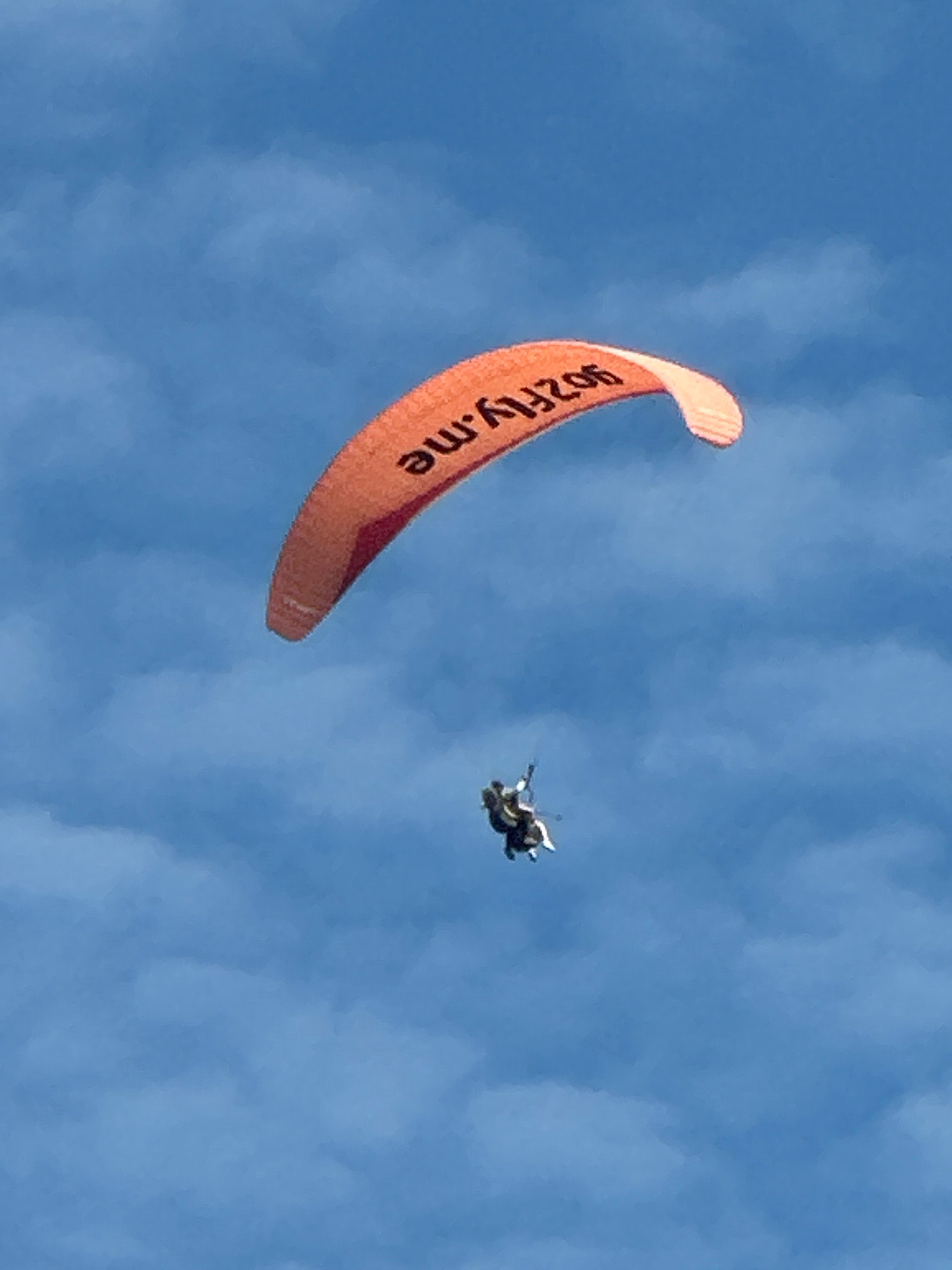 An orange paraglider with the black text "902FLY.ME" written on it is soaring through a blue sky with scattered white clouds. Beneath the paraglider, two individuals are tandem paragliding, suspended in the air with their legs dangling. The scene appears to be a recreational activity, possibly in the vicinity of Boreti, Montenegro, known for its coastal beauty and adventure tourism. The overall impression is one of freedom and exhilaration against a backdrop of nature's elements.