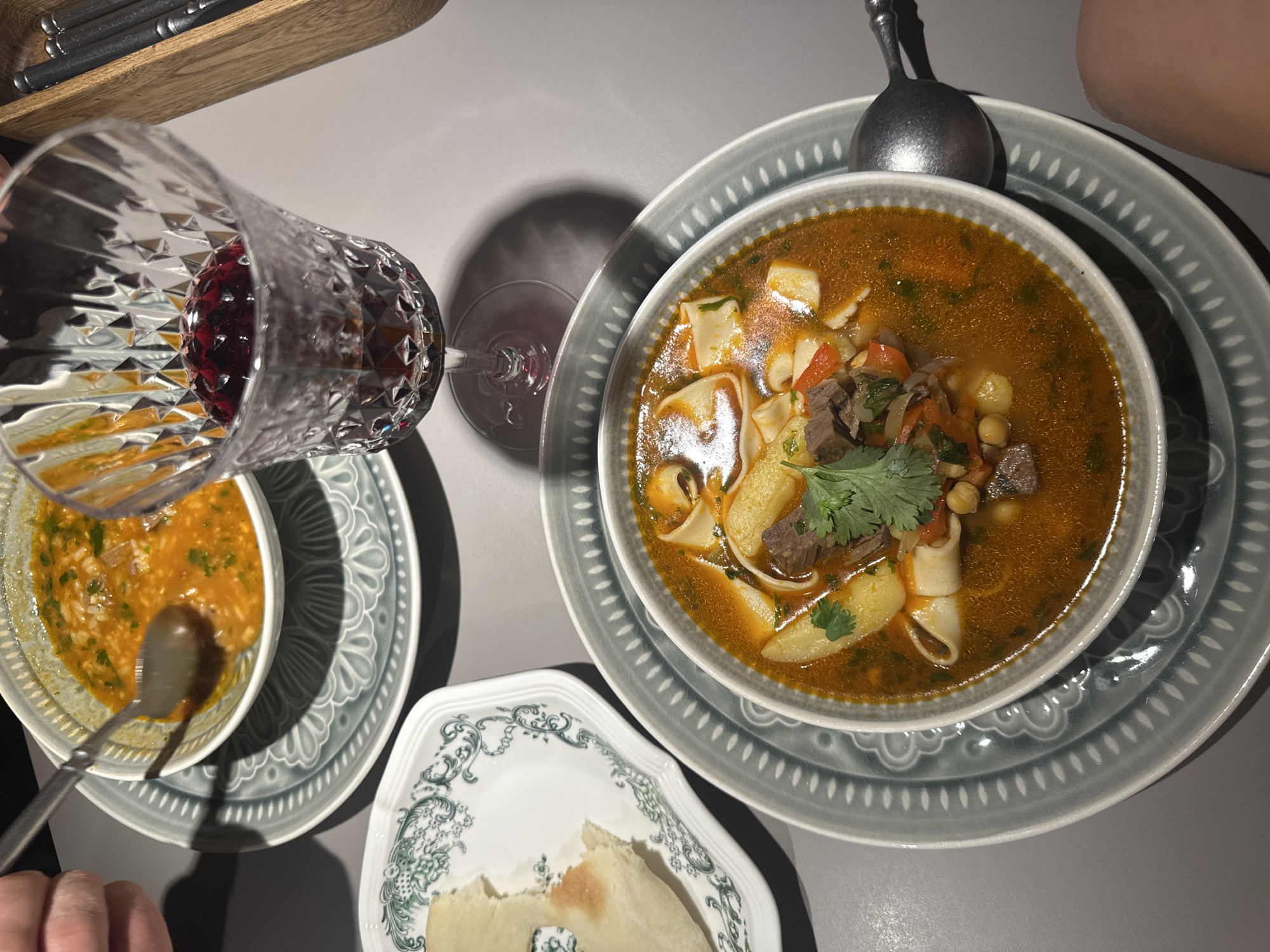 The image depicts an overhead view of a dining table in Chaikovsky, Russia, set for a meal involving two individuals.
In the foreground, a large, light grey ceramic bowl containing a hearty, orange-red soup is centered on a larger, circular grey ceramic plate with an embossed floral and scalloped rim design. The soup contains thick, flat noodles, cubed pieces of dark meat (beef or lamb), cubed yellow potatoes, chickpeas, diced red vegetables (possibly carrots or bell peppers), and is garnished with fresh green cilantro. A silver spoon with an ornate handle rests in the soup, pointing towards the lower right.
To the upper left, a second, smaller light grey ceramic bowl, also on a matching patterned plate, holds a lighter orange-colored soup containing grains (likely rice or small pasta) and green herbs. A hand with a light skin tone, wearing a long-sleeved garment, holds a silver spoon partially submerged in this soup, indicating active consumption.
Between the two soup bowls, slightly to the right, a stemmed, clear crystal glass with a faceted pattern holds a dark red liquid, approximately one-third full, likely red wine or a berry beverage. Below the glass, its shadow is cast on the table.
To the mid-left, a rectangular white ceramic plate with a green decorative floral pattern on its rim holds a piece of light-colored, torn flatbread.
In the upper right corner, a light-colored wooden rectangular container holds several dark, slender utensils, possibly chopsticks.
The table surface is a smooth, matte light grey. A bare knee or thigh with a light skin tone is visible at the very bottom right of the frame, indicating the presence of a second diner. The lighting is uniform, suggesting an indoor setting.