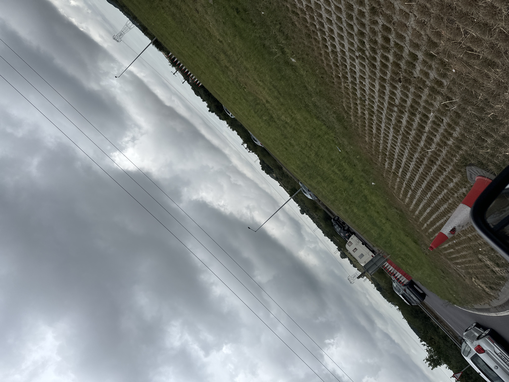 The image, taken from a low angle, depicts a traffic scene on a cloudy day in an unspecified location in Ireland. The foreground is dominated by a textured, honeycomb-like ground cover, likely a permeable paving system, with sparse dry grass. A portion of a vehicle's side mirror, tinted dark, is visible on the left. A red and white traffic cone is positioned on the ground.
The middle ground shows a grassy verge bordering a road where several vehicles are present, appearing to be in slow-moving traffic or stopped. These include a silver Volkswagen, a dark-colored SUV, a white caravan or trailer being towed, and at least two other dark passenger cars. Orange and white traffic cones line a section of the road. Further back, a dense line of trees forms the horizon, with a power line pylon and streetlights visible against the overcast sky. The sky itself is filled with heavy, grey, dramatic clouds. The overall scene suggests a roadside environment, possibly an approach to or exit from a town or city, or a section of roadworks.