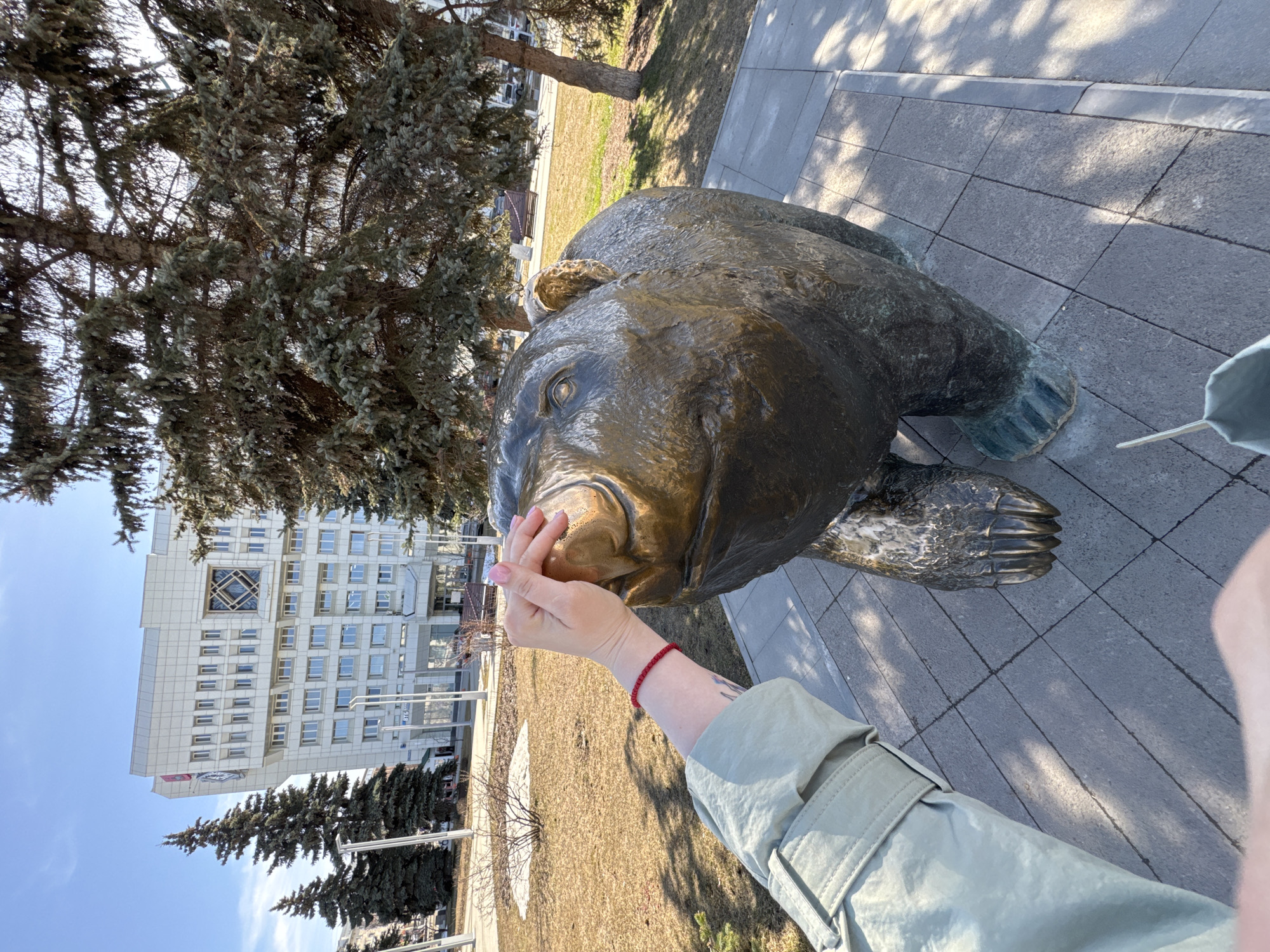 The image shows a bronze bear statue in the city of Perm, Russia, near the Permyak Salted Ears monument, located in front of the Perm-1 railway station. A person is touching the statue's nose with their fingers. The person wears a light green jacket and a red bracelet is on their left wrist. A large building is visible in the background on the left side of the image. There are trees nearby and a paved area surrounds the statue.