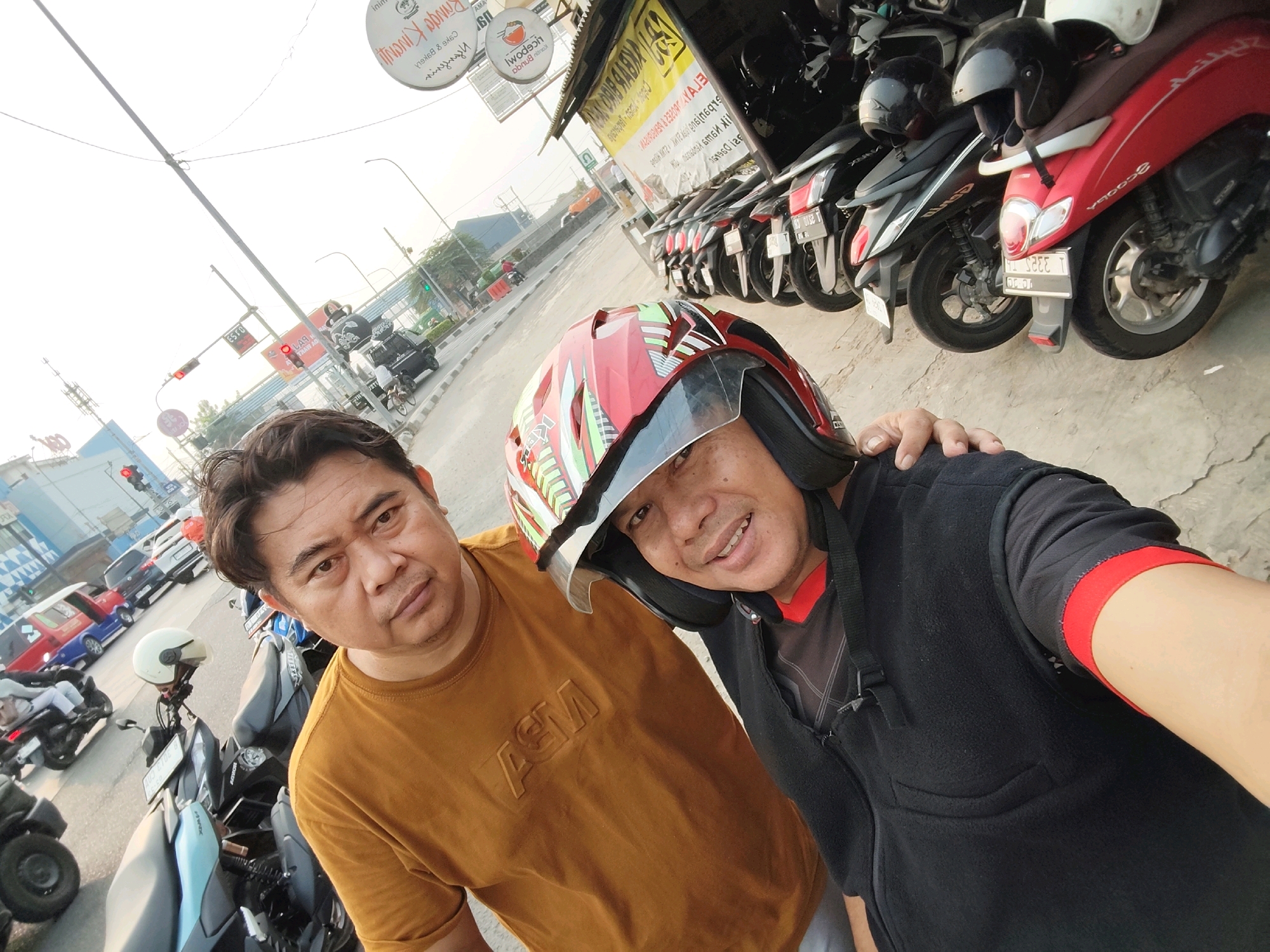 Two Indonesian men are taking a selfie outdoors in what appears to be a commercial area in Purwakarta, Indonesia. The man in the foreground on the right is wearing a red and green patterned helmet with the visor up, a black vest over a black and red patterned shirt, and a black strap across his chest. He is smiling. The man on the left is wearing a mustard yellow t-shirt with a raised "ASM" logo and has his arm around the man on the right. Both men are looking at the camera.
In the background, there is a row of parked motorcycles, including a red scooter on the far right and several black scooters. A street with traffic is visible behind the motorcycles. There are also buildings and signs, including one with text in Indonesian that appears to be advertising food or drinks. The overall lighting suggests it is daytime, possibly late afternoon due to the warm light and long shadows. The scene is consistent with a public space in an urban environment in Indonesia.