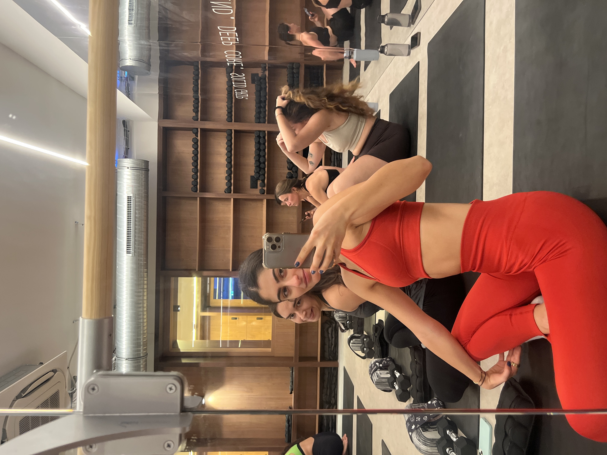 The image depicts a group of women in a fitness studio, likely for a workout session. The women are dressed in athletic wear and are positioned on yoga mats. One woman in the foreground is taking a selfie with her phone, wearing a red sports bra and leggings. Another woman next to her is looking at the phone. In the background, other women are engaged in exercises or resting. The studio has wooden shelves filled with dumbbells, and ventilation pipes and lighting fixtures are visible on the ceiling. The overall environment suggests a clean, modern gym or studio setting. There is no indication of the specific location within Paris, France.