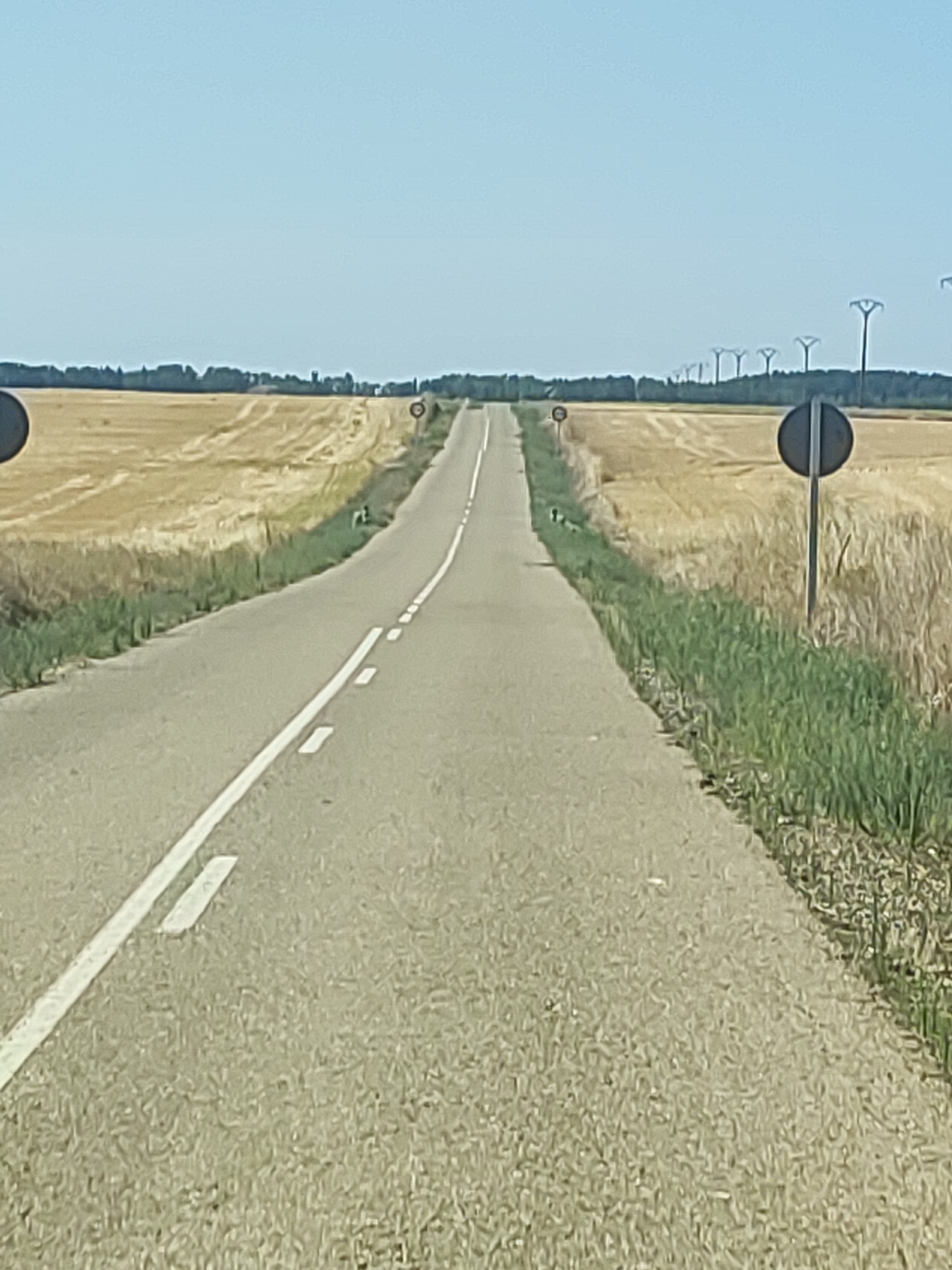 The image shows an empty asphalt road with white dashed lines and a solid white line marking the lane. The road is surrounded by dry, golden fields of harvested crops. In the distance, a line of tall, Y-shaped pylons stretches across the horizon, silhouetted against a clear blue sky. Two black, circular traffic signs are visible on poles along the side of the road. One sign is in the foreground on the right, and another is further down the road on the left. The environment suggests a rural, agricultural setting. The absence of people or vehicles indicates a quiet, undeveloped area. This scene is consistent with the landscape of Castromocho, Spain, a municipality known for its agricultural land and open fields.