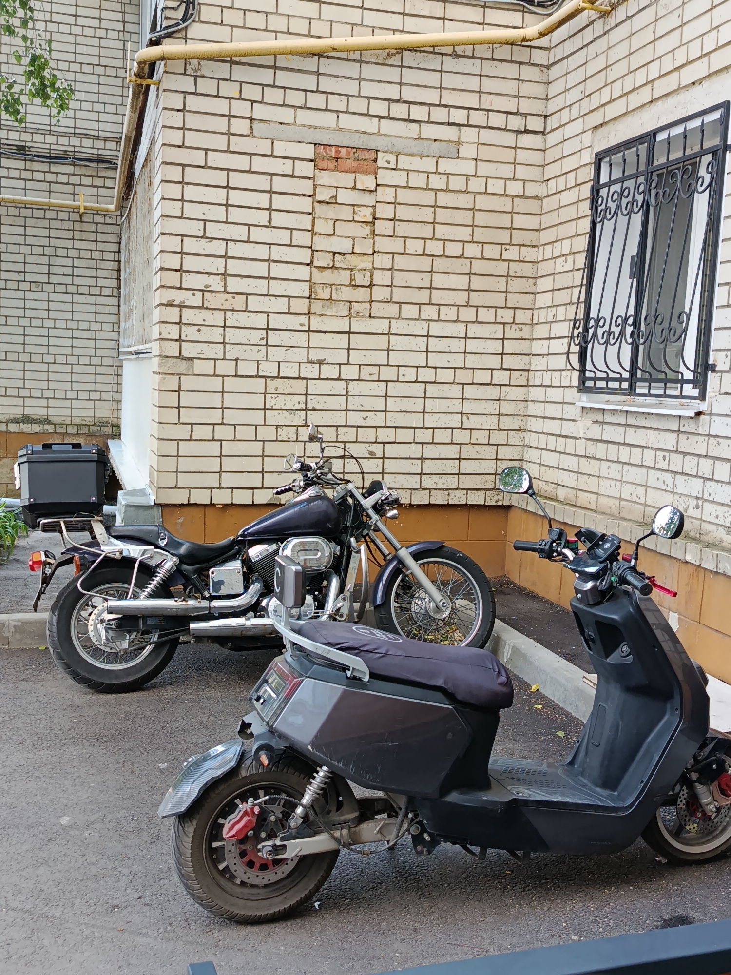 Two parked vehicles, a dark colored scooter in the foreground and a dark blue motorcycle in the background, are situated on an asphalt surface next to a beige brick building. The building has a yellow pipe running along its exterior and a barred window. The scooter has a gray seat cover and a carbon fiber fender over its rear wheel, which features a red brake caliper. The motorcycle has a black luggage box mounted on its rear rack, chrome exhaust pipes, and a spoked front wheel. The scene appears to be an outdoor urban environment.