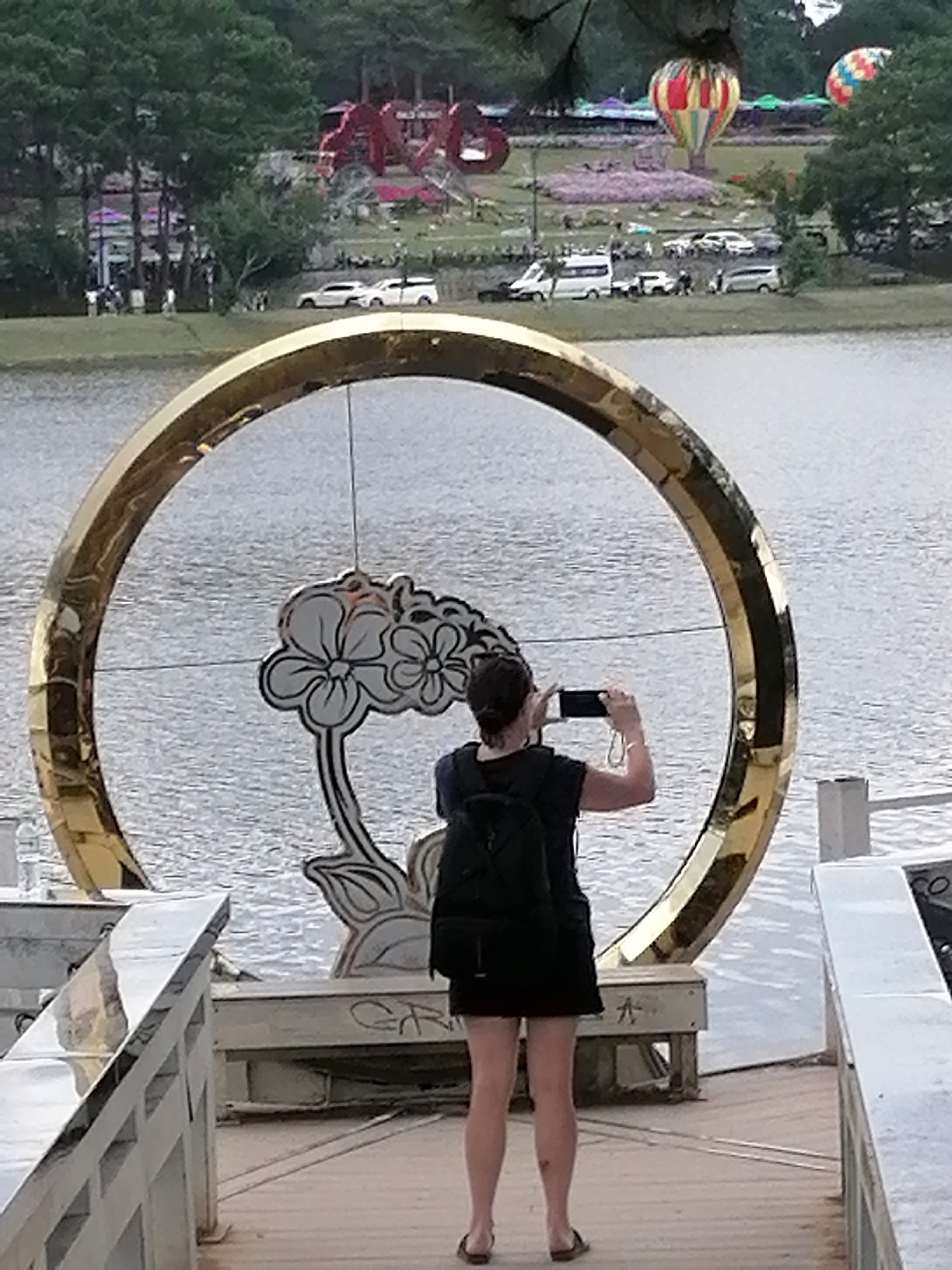 A woman with a black backpack stands on a wooden walkway, facing away from the camera, taking a picture of a large golden ring structure with her phone. The ring features a stylized floral design. Behind the ring, a body of water is visible, with a distant park containing trees, flowers, vehicles, and hot air balloons. The scene appears to be in Da Lat, Vietnam, likely near Xuan Huong Lake, a popular tourist attraction known for its hot air balloon festivals and parkland.