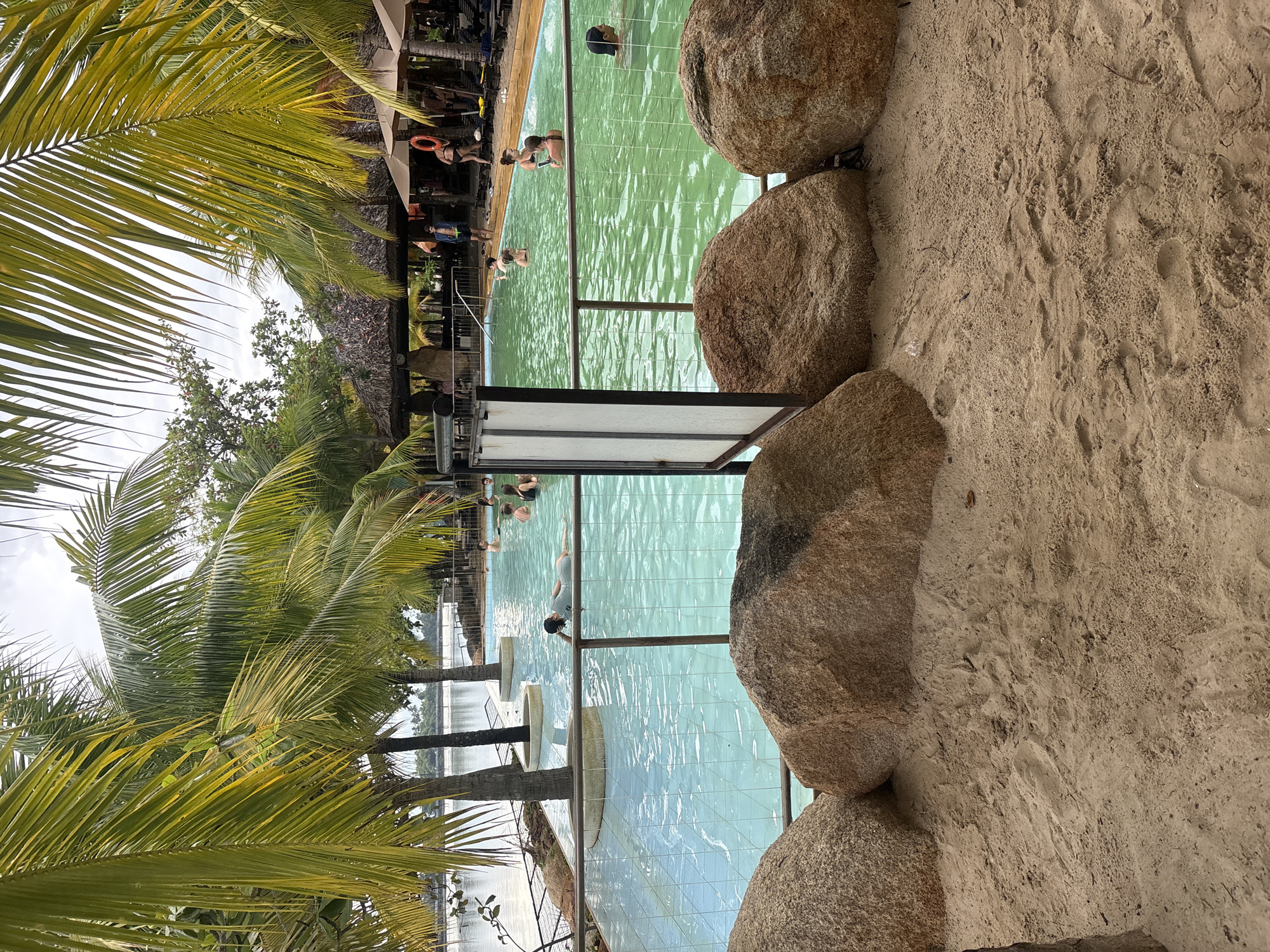 This image depicts a resort swimming pool area in Nha Trang, Vietnam, characterized by palm trees, a sandy foreground with large rocks, and a pool filled with people. The pool itself is a prominent feature, with light blue tiles visible in some sections and a greenish hue in others. Several individuals are swimming and relaxing in the water. A thatched-roof structure with umbrellas and lounge chairs suggests amenities for guests. The foreground is composed of sand with visible footprints and embedded rocks, indicating a beach-like environment adjacent to the pool. The presence of palm fronds in the upper part of the image reinforces the tropical setting. The sky is overcast, suggesting a diffused light condition. The overall scene captures a leisure activity in a resort setting.