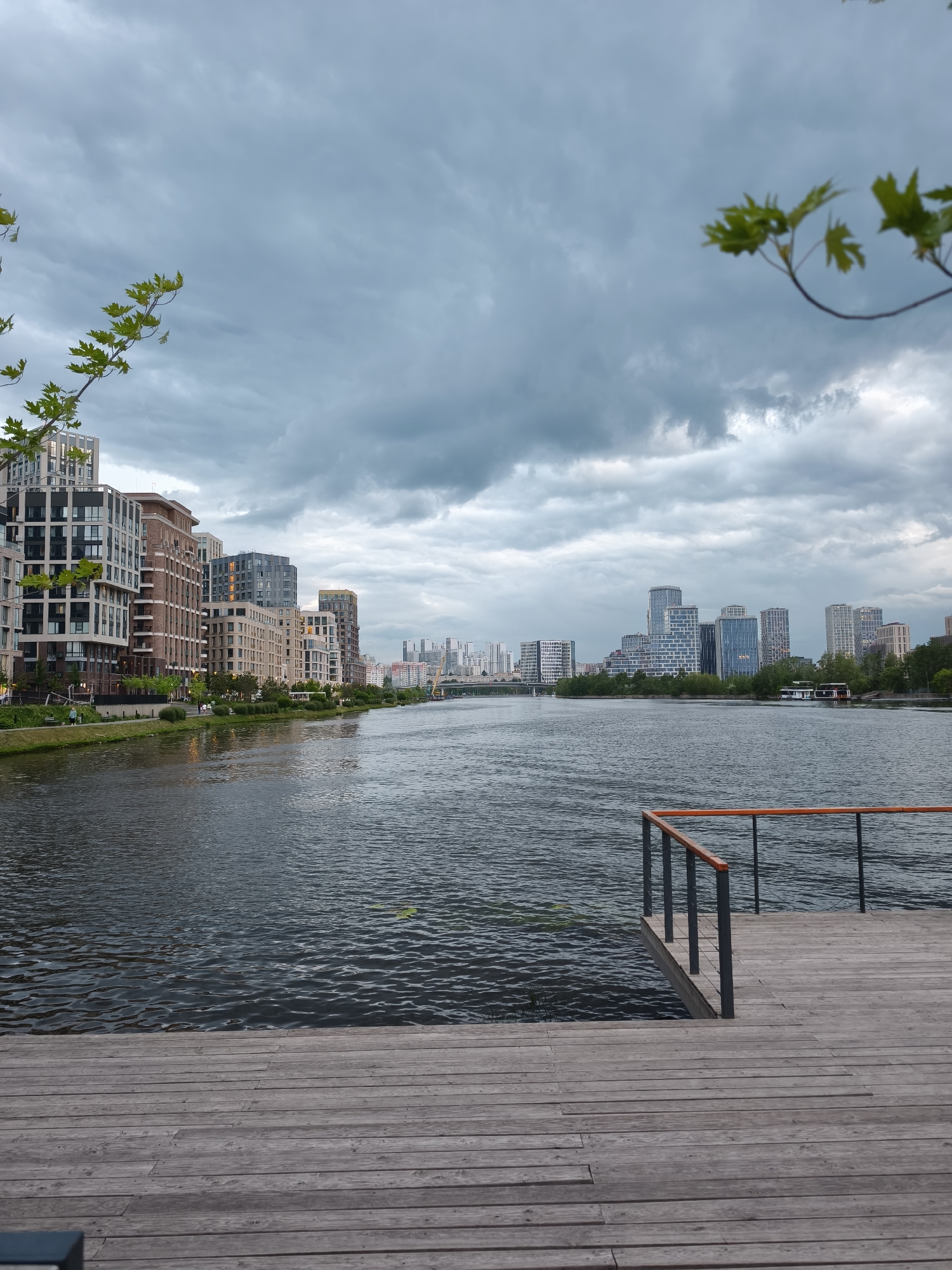The image captures a waterfront scene in Moscow, Russia, under an overcast sky with heavy, grey clouds. The perspective is from a wooden deck or pier with a grey metal and wood railing extending into the water. The water appears calm with gentle ripples.
Across the water to the left is a row of modern residential buildings of varying heights and architectural styles, set against a green strip of grass and trees along the riverbank. Further in the distance, across the water, more modern high-rise buildings are visible.
Green foliage is partially visible in the top left and top right corners of the frame, suggesting trees near the viewing point. No people are visible in the image.