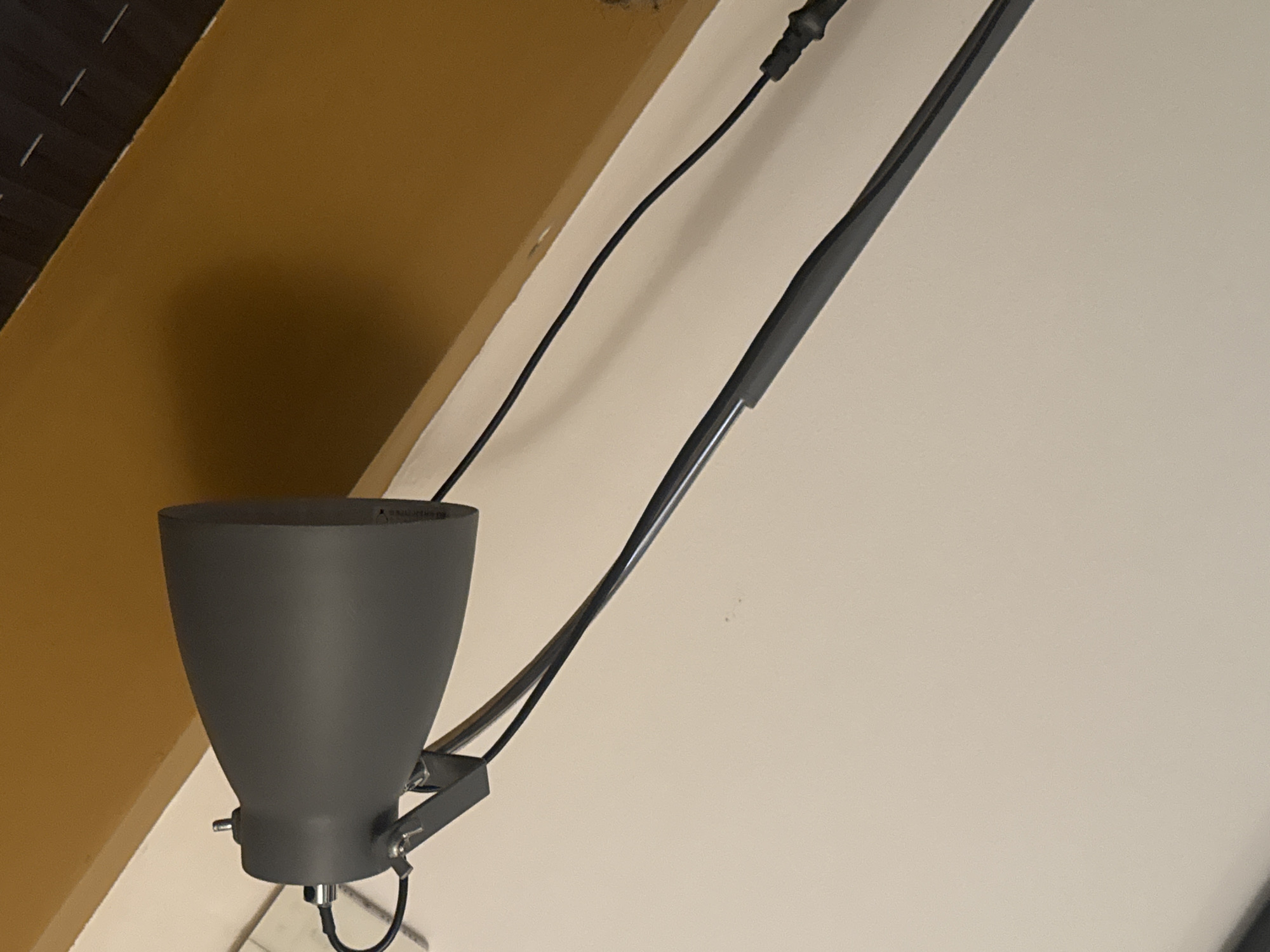 A dark grey, adjustable desk lamp with a conical shade is positioned on a light-colored, textured surface, possibly a wall or a desk. The lamp's flexible arm extends downwards and to the right, with a black cord trailing away from the base. In the background, a horizontal band of warm, golden-brown color is visible, possibly indicating a wall or ceiling with a different finish. The lighting suggests an indoor environment, and the overall impression is one of a functional, modern object. The image does not provide enough detail to determine its specific location in a city.