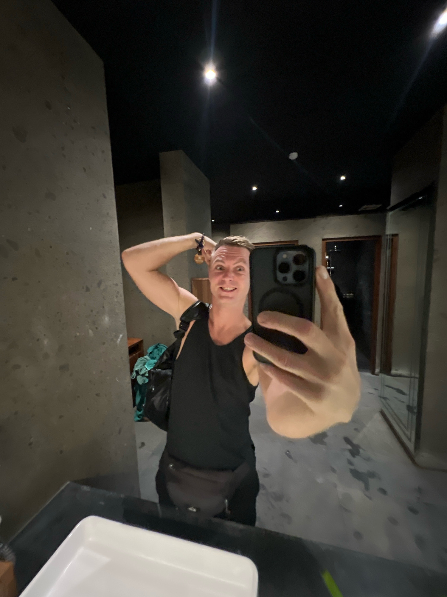 A man is taking a selfie in a dimly lit, modern restroom. He is wearing a black tank top and a fanny pack. He has his right hand on his head and is holding a smartphone in his left hand, which is angled towards the mirror. The restroom has concrete walls and a dark ceiling with several recessed lights. A white sink is visible in the foreground. In the reflection, a black backpack with a teal item inside is on the floor behind the man. The floor appears wet in places. The location is described as being in the Ubud District, Indonesia, which suggests a tropical, possibly humid, environment.