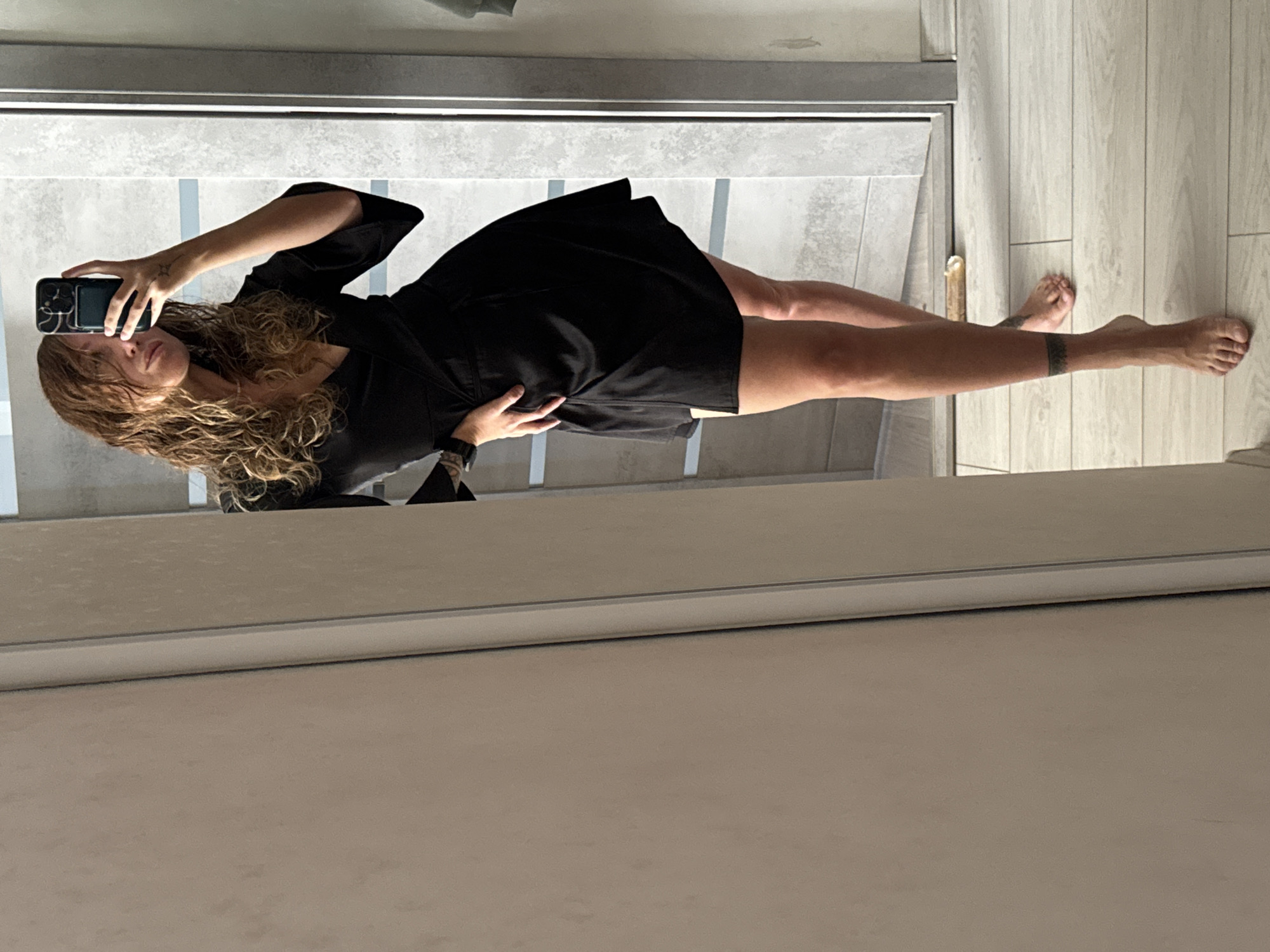 A woman is taking a mirror selfie. She is wearing a black dress. She is holding a cell phone in her left hand. She has curly blonde hair. She is standing on a light-colored wooden floor. The location cannot be verified based on the image provided.