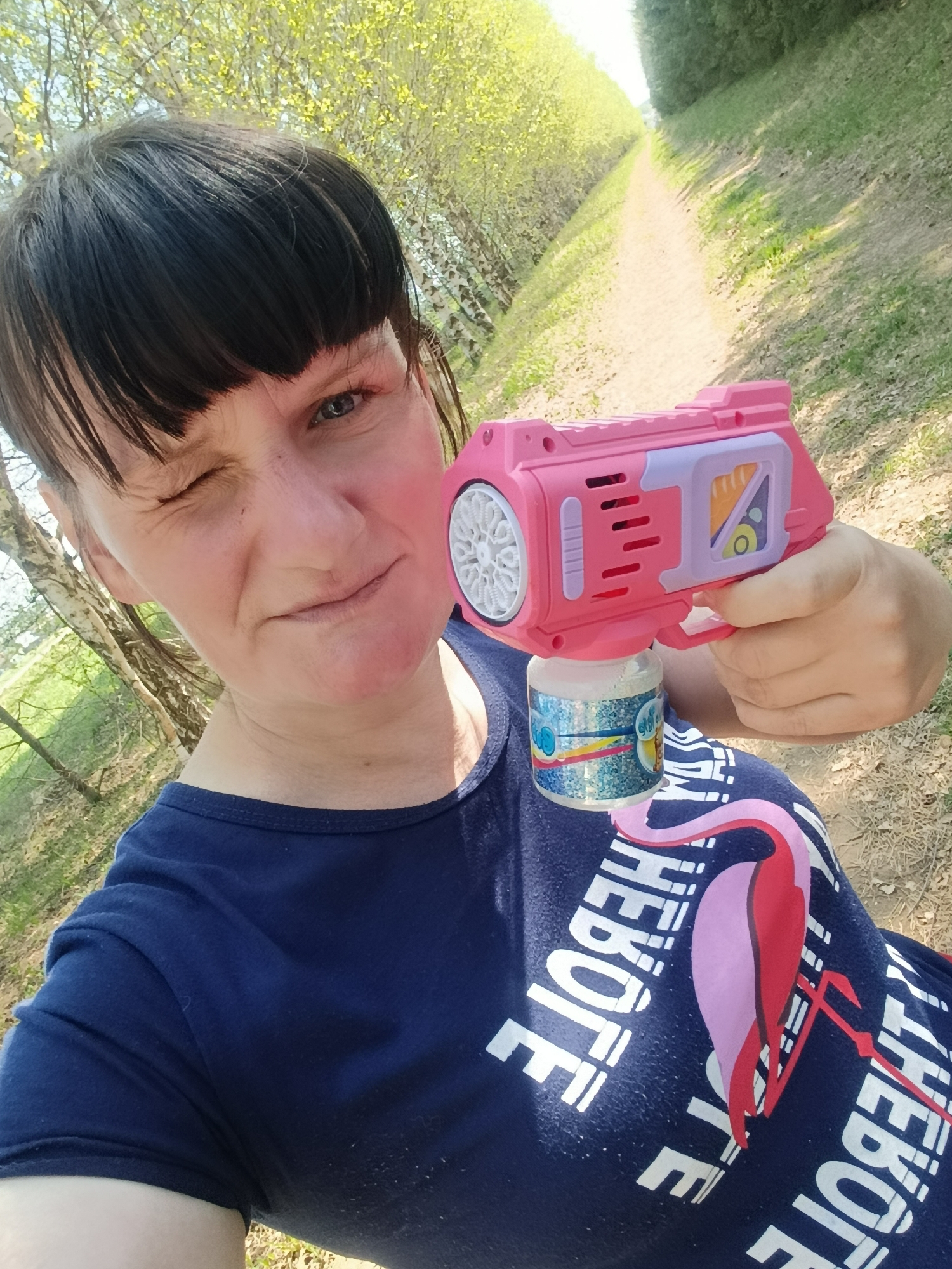 A medium shot depicts a woman outdoors holding a pink bubble gun. She is partially winking at the camera. She has short, dark hair with bangs and is wearing a dark blue t-shirt with a flamingo graphic and text. The bubble gun is pink with a white bubble-blowing wheel and a bottle of bubble solution attached to the bottom. The background shows a dirt path lined with trees and foliage, suggesting a park or outdoor recreational area. The specific location is in the city of Фоки, Russia.