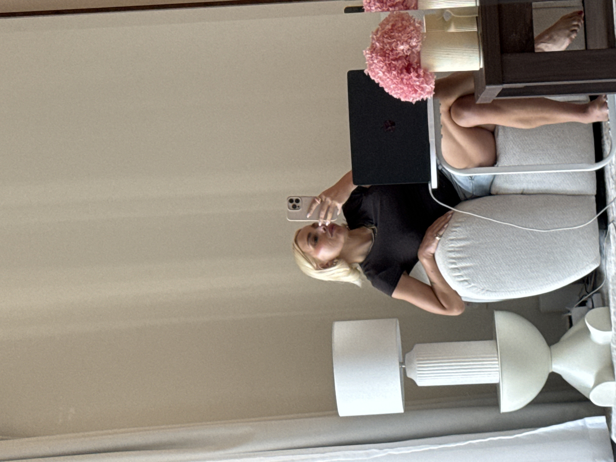 The image displays a mirror selfie of a blonde woman taken in an indoor setting, possibly a hotel room or apartment in Dubai. The woman, positioned centrally, holds a light-colored smartphone to capture her reflection. She is wearing a black short-sleeved top and light-colored bottoms, with a ring visible on her right hand. Her expression is neutral to slightly smiling as she looks at the phone.
She is seated in a comfortable, light-colored armchair with plush cushions. To her immediate left, a black laptop (possibly a MacBook given the discernible logo) rests on a surface, partially obscured by a bright pink floral arrangement. Her legs are visible to the right, casually crossed. Further right, a dark wooden side table or furniture piece holds a bare foot with red painted toenails.
The room's decor is modern and minimalist, featuring light-colored walls and a soft, ambient light, suggesting it is daytime. Two distinct lamps are visible: one with a white cylindrical shade and a ribbed base, and another with a sculptural white base topped by a half-sphere. The overall atmosphere is relaxed and casual. There is no discernible text visible in the image.