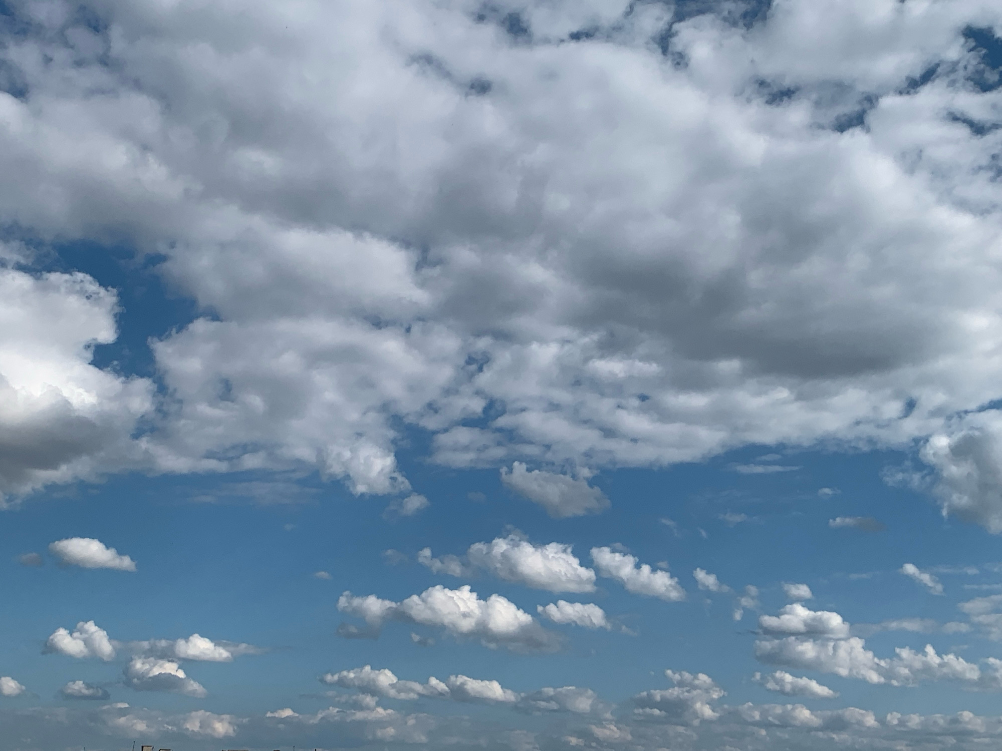 The image displays a sky filled with a mix of fluffy white and darker gray clouds against a clear blue sky. The clouds are scattered, with some large formations dominating the upper portion of the frame and smaller, more distinct clouds lower down. The lighting suggests it is daytime, with sunlight illuminating the tops of the clouds. The bottom edge of the image shows a faint hint of a distant landscape, possibly the outskirts of a city, with the suggestion of structures, but no clear details are discernible. The image is a general sky view and does not contain any specific landmarks or elements that would pinpoint a location within Kolomyia, Ukraine, or any other city. There are no people, objects, or actions depicted in the image.