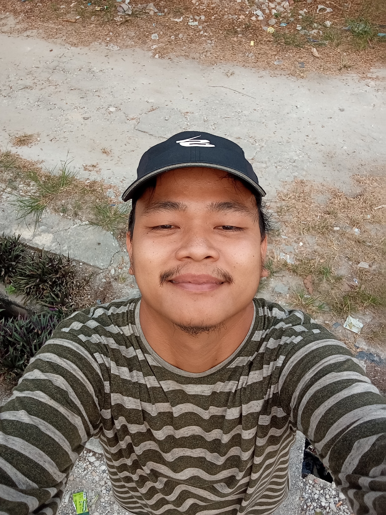 A young male wearing a black baseball cap and a striped long-sleeved shirt is captured in a selfie outdoors. The background consists of a dirt path with sparse dry grass, some gravel, and a patch of dark green leafy plants. The man has a mustache and a goatee. The scene appears to be in an outdoor area, possibly a park or roadside in Kualu, Indonesia.