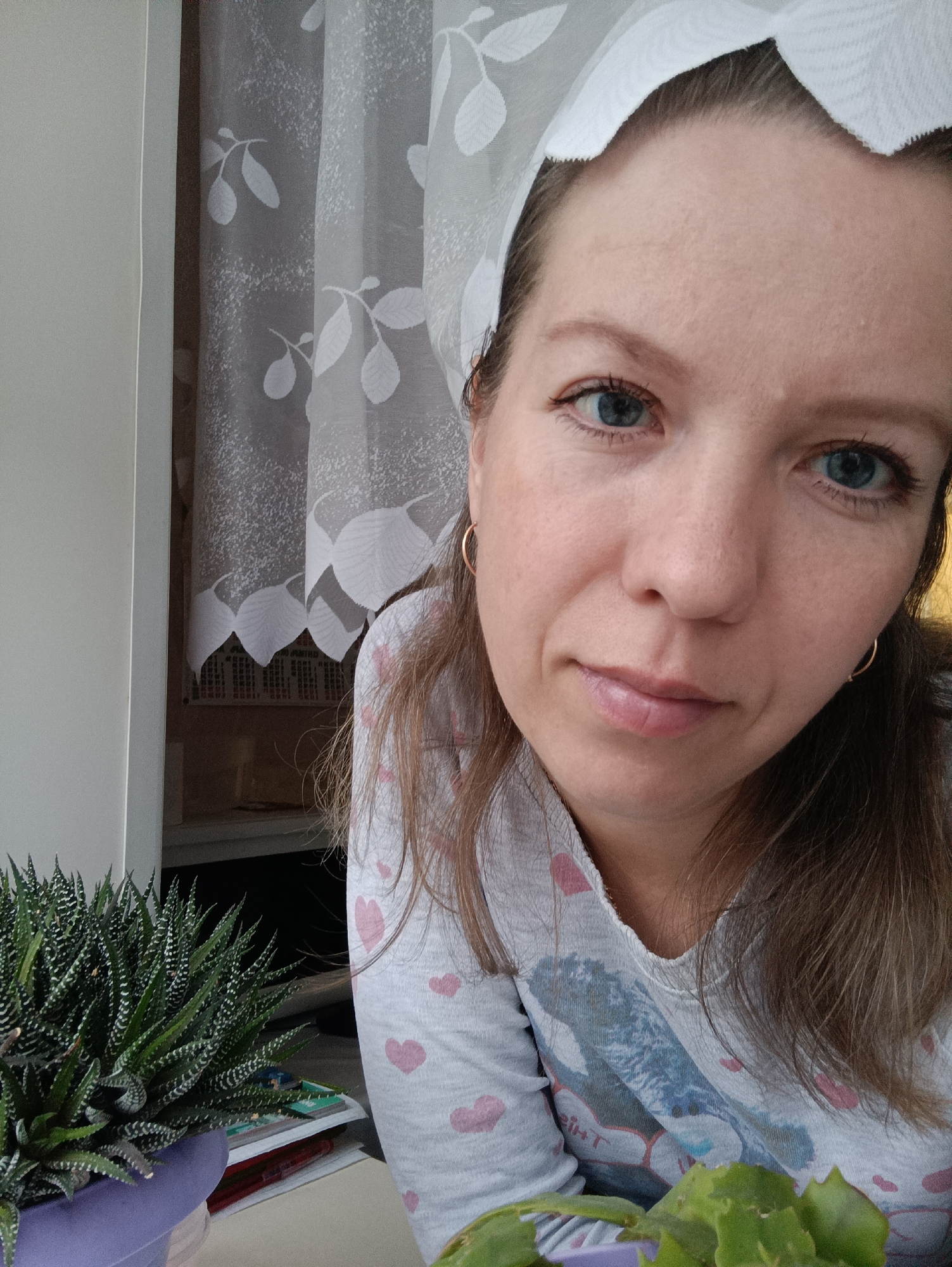 A woman is indoors in what appears to be a room in Chaikovsky, Russia, based on available information. She is close to a window covered by a white curtain with a floral pattern. A potted succulent plant with striped green leaves is to her left. The woman has blue eyes, light brown hair, and wears gold hoop earrings. She is wearing a light gray top with pink hearts and a blue print on the front. A piece of white lace fabric is on her head. The photo appears to have been taken during the day with natural lighting.