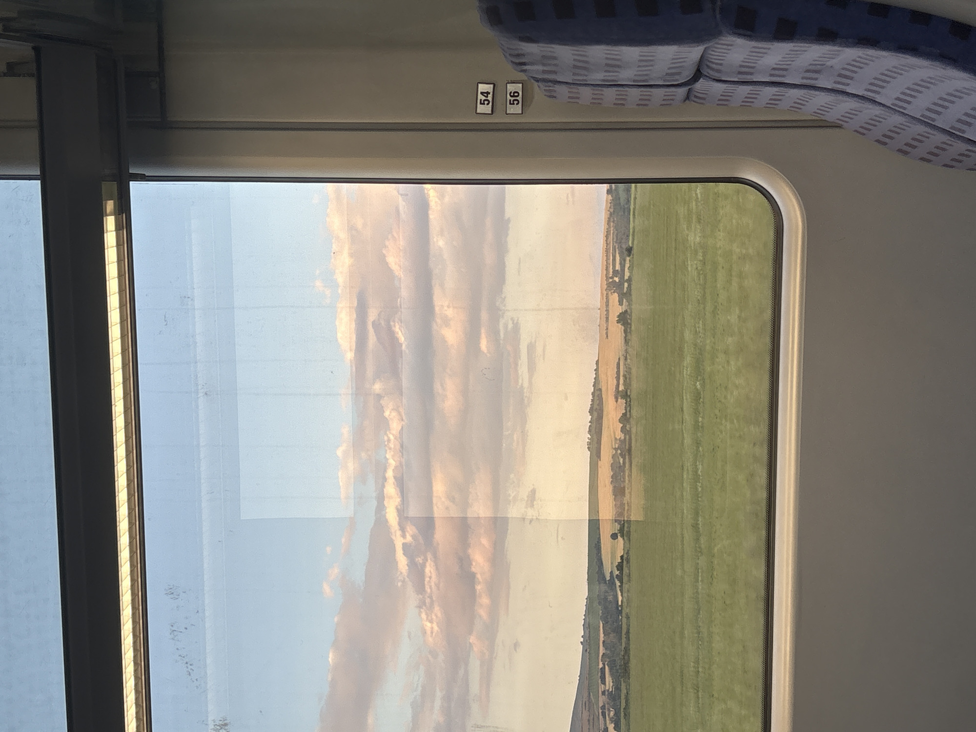 This image depicts a view from inside a train looking out a window at a rural landscape under a partially cloudy sky, with the provided context placing the scene in the city of Svoboda, Bulgaria. On the right side of the frame, a segment of a train seat is visible, upholstered in a blue fabric with a repeating pattern of light-colored, thin horizontal and vertical lines. Above the seat, two white rectangular labels with black numbering, "54" and "56", are affixed vertically to the light-colored interior wall of the train. A light fixture, featuring a grid-patterned diffuser emitting a warm yellow glow, runs horizontally above the window frame. The interior window frame is light grey with a rounded bottom edge. Faint reflections of the train's interior are visible on the window glass, including a prominent rectangular light area on the right and scattered smudges or dust particles on the upper left pane.
Through the window, the sky displays a gradient of color, from light blue at the top to soft pink and orange hues near the horizon, characteristic of twilight. Multiple altocumulus or stratocumulus clouds, primarily white with subtle pink and orange tints, are distributed across the sky. The landscape below the sky consists of agricultural fields and rolling terrain. The immediate foreground is dominated by a wide expanse of vivid green crops. Beyond this green field, the midground features a mosaic of golden-yellow fields, likely mature grain, intermingled with clusters of dark green trees and bushes. A large, verdant hill or low mountain is discernible on the far left of the horizon. The entire external scene is illuminated by soft, warm, natural light.