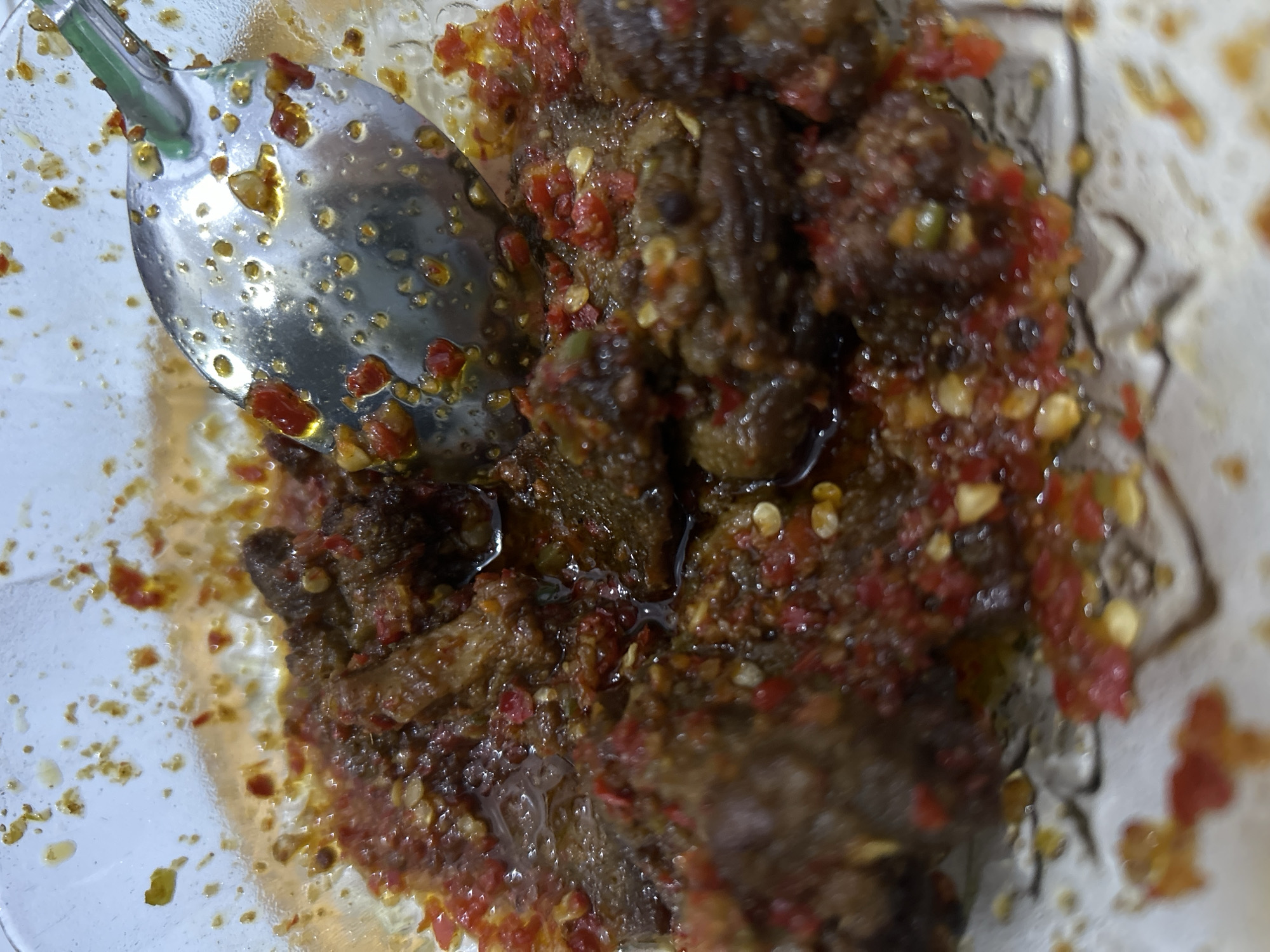 A close-up shot depicts a dish with pieces of dark meat covered in a red chili-based sauce, possibly chili paste or sambal. A metal spoon rests on the side of the dish. The dish is in a clear glass bowl. Based on the information given, the photograph was taken in Lhokseumawe, Indonesia.