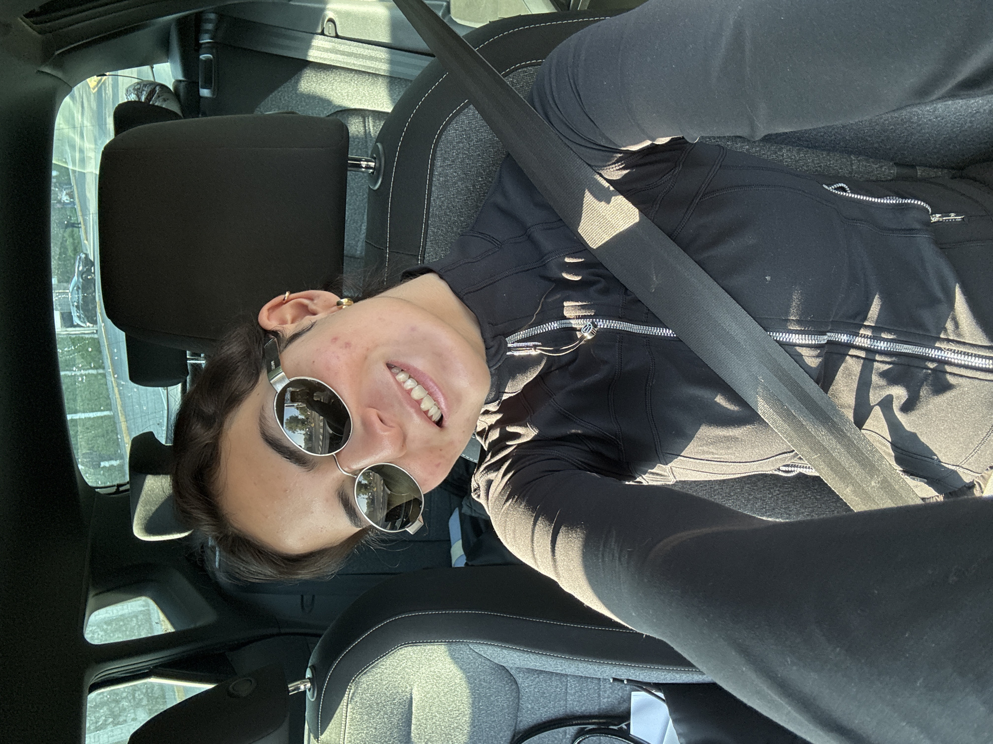 The image is a selfie taken inside a car. A young woman wearing round sunglasses and a black athletic jacket is facing the camera, with a seatbelt across her chest. The interior of the car is visible, including the seats and headrests. Through the car window, there is a view of a cityscape, possibly in the city of Puebla, Mexico, based on the architectural styles and street patterns visible in the reflection.