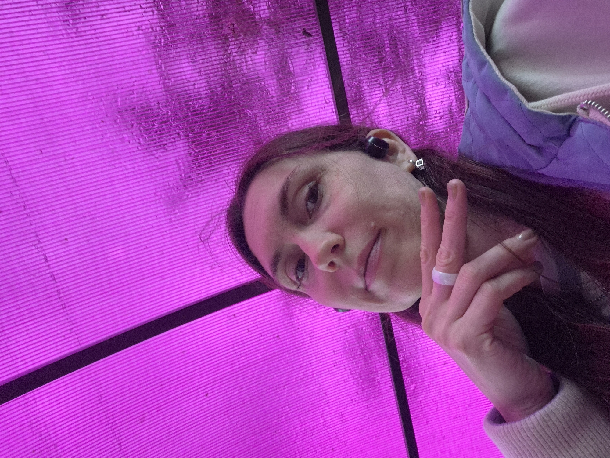 A woman poses for a selfie in a location with bright pink illumination. She is wearing a ring on her right hand and earbud in her left ear, and is giving a peace sign. Part of a jacket can be seen on her right shoulder. The background has linear patterns, appearing to be a wall or ceiling structure with pink lighting. No information from the image indicates it being in the city of Chaikovsky, Russia.
