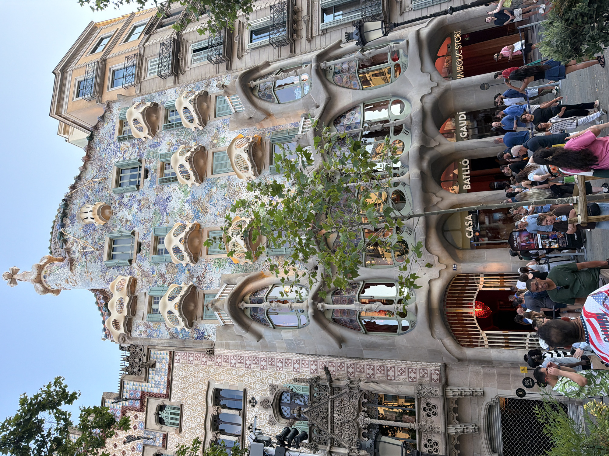 The image depicts Casa Batlló, a landmark building designed by Antoni Gaudí, located on Passeig de Gràcia in Barcelona, Spain. The ornate facade of the building is covered in colorful mosaic tiles and features undulating balconies that resemble bones and masks. Several trees with green foliage are in front of the building, partially obscuring the lower floors. A crowd of people is gathered on the sidewalk, many of whom appear to be tourists. Some are looking at the building, while others are walking or standing in groups. The sky is clear and blue, indicating it is daytime. Signs for "CASA BATLLÓ" and "GAUDI" are visible on the building's entrance.