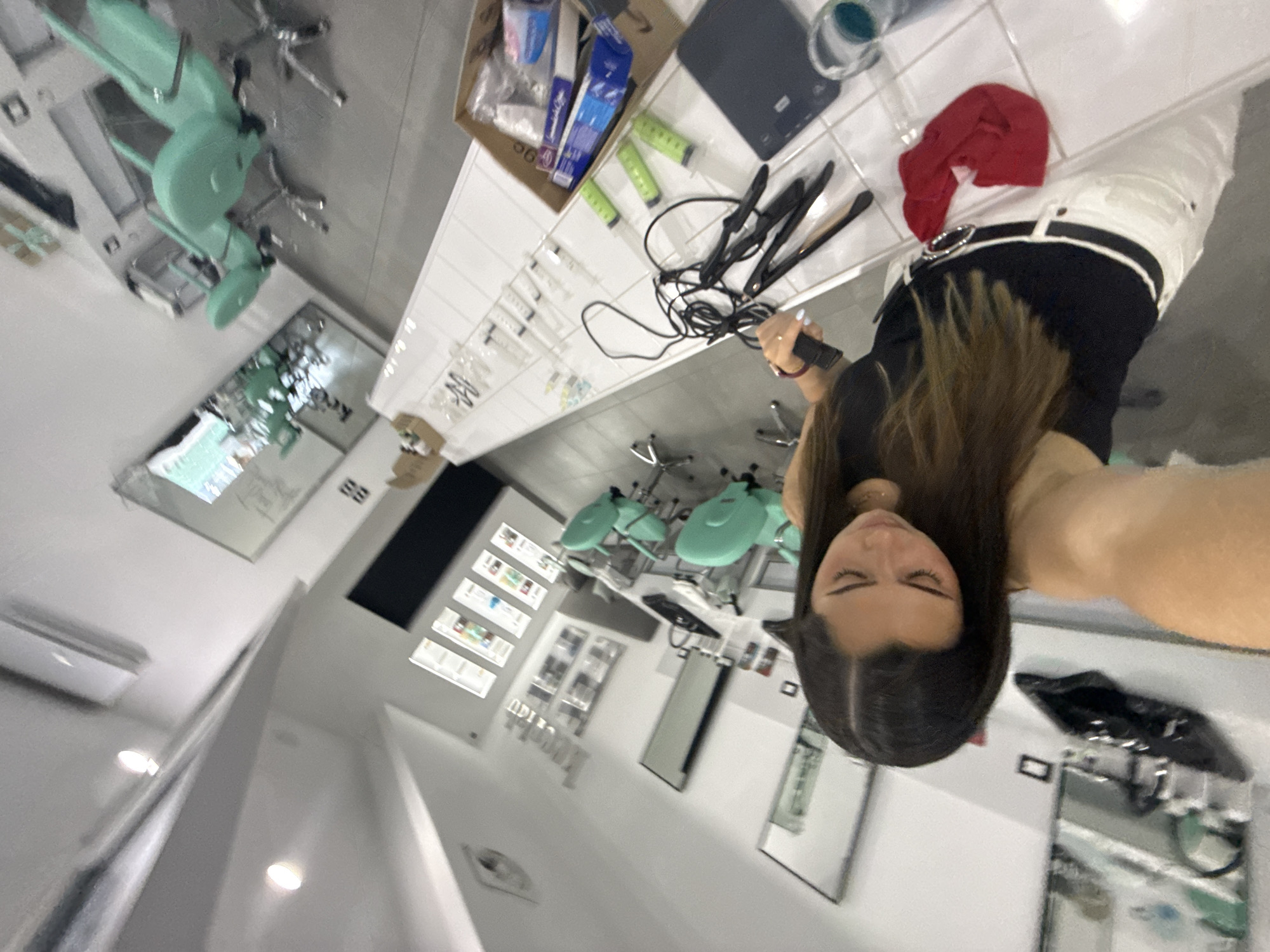 The photograph shows the interior of what appears to be a beauty salon or aesthetic clinic. A young woman is holding a curling iron. The salon has multiple treatment chairs, a display of products, and white tiled surfaces. Medical supplies, including syringes, are visible on the table. The location is presumably San Bernardino Tlaxcalancingo, Mexico.