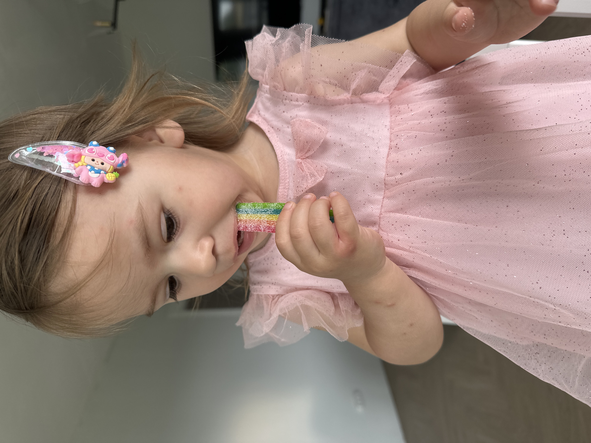 A young child with light brown hair is shown in an indoor setting, wearing a pink, sparkly tulle dress. A hair clip featuring a cartoon character is visible in the child's hair. The child is holding and eating a rainbow-colored candy strip. The exact location within the city of Kharnavy, Russia cannot be determined.