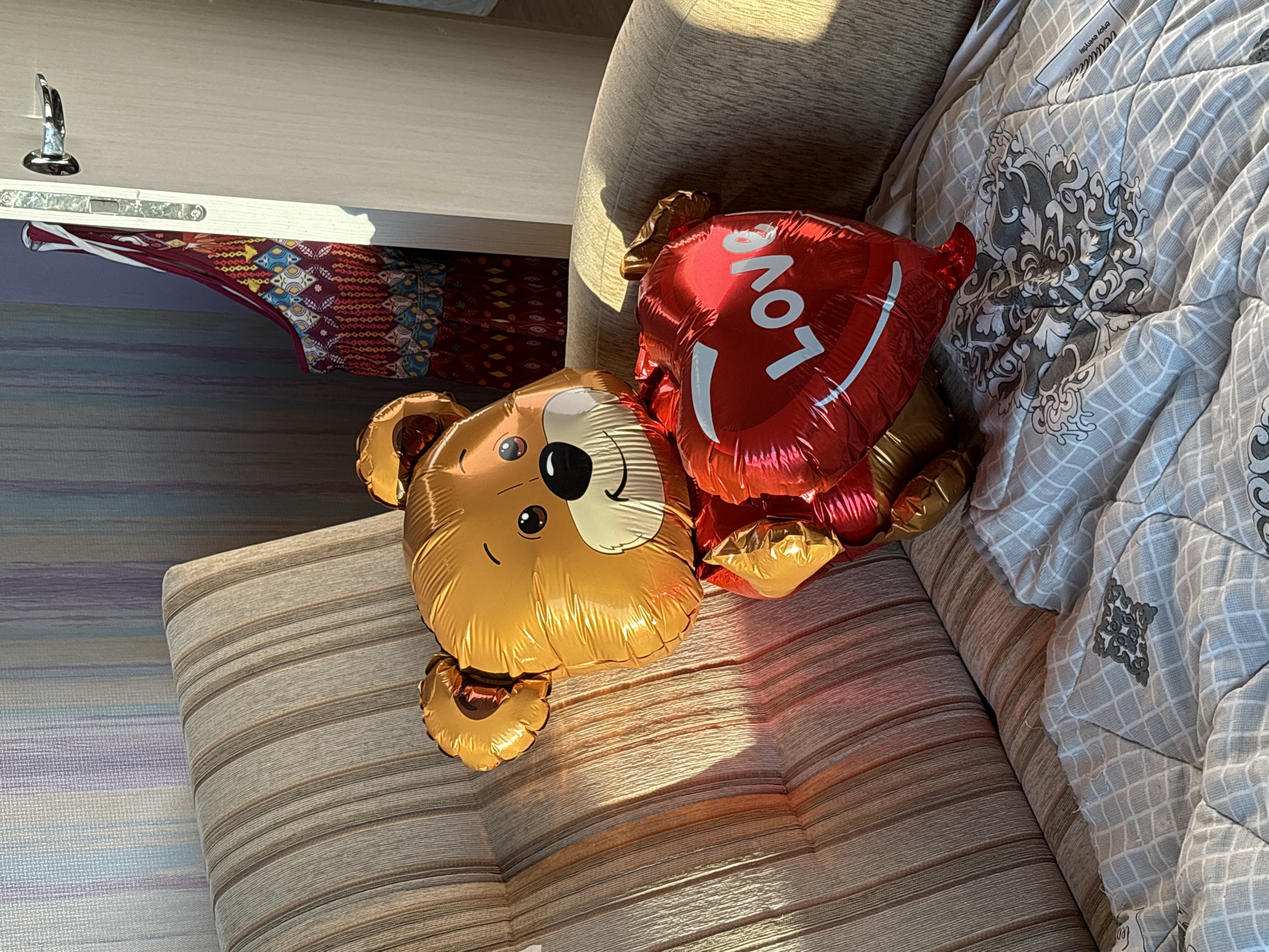 A teddy bear balloon, gold in color with a red heart balloon that reads "Love," sits on a couch in an interior room. The couch is upholstered in a striped fabric of tan and brown. The balloon is positioned on the seat cushion. Behind the couch, a door with a silver handle is slightly ajar, revealing a colorful garment hanging inside. The wall behind the couch is decorated with vertical stripes of pale blue and lavender. The floor is covered with a light-colored blanket featuring a repeating grid pattern and a decorative medallion in gray. Sunlight streams into the room from the right, casting shadows on the couch and floor. There are no people present in the image. The location is identified as Novoselye, Russia.