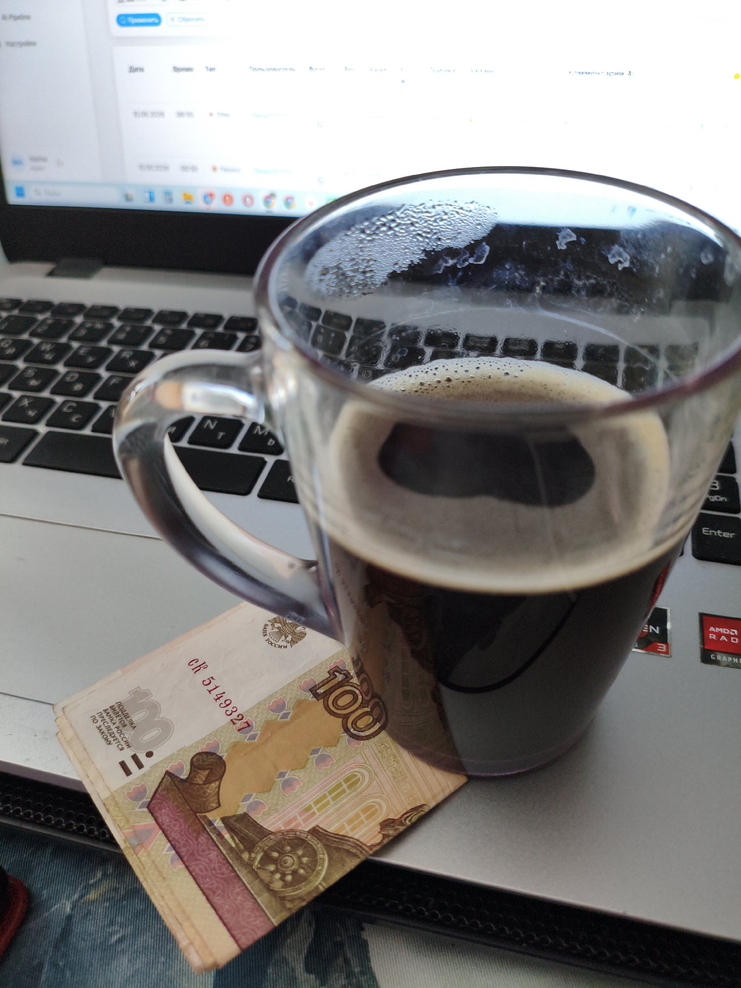 The image shows a clear glass mug filled with a dark liquid, likely coffee, placed on a laptop. Condensation is visible on the outside of the mug. A few Russian ruble banknotes, specifically one hundred ruble notes, are folded and tucked under the mug. The laptop's keyboard and screen are partially visible. The screen displays what appears to be a web page or application with text and icons. The overall scene suggests a work or study setting, with the coffee and money implying a break or a business-related context. The image does not provide any specific information about the city of Orenburg, Russia.