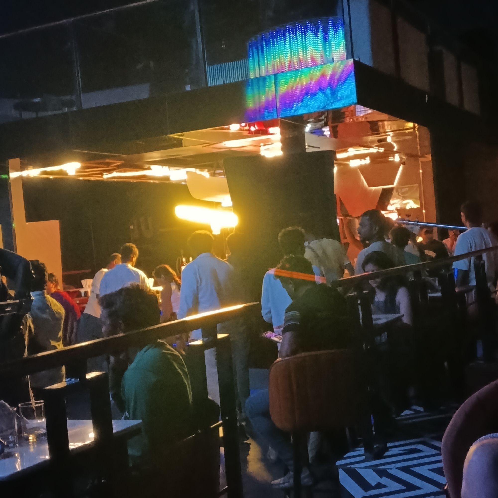 A crowded indoor scene, likely a bar or nightclub, is captured at night in Sinquerim, India. Several people are seen standing and sitting, dressed in casual attire. Light is predominantly yellow-orange, with a digital display exhibiting a rainbow pattern overhead. Tables with drinks are visible in the foreground. A dark railing separates foreground from background. The floor has a patterned design.