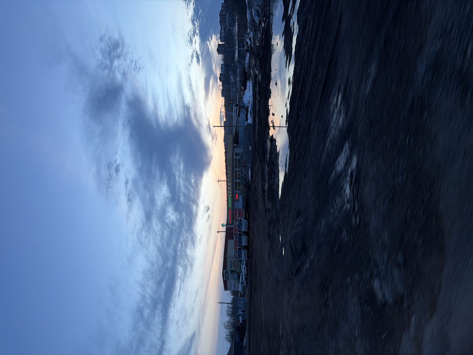 The image shows a street scene in Votkinsk, Russia, at dusk or dawn. The sky is a mix of blue and orange hues with scattered clouds. On one side of the image, there's a row of buildings, with some signage and parked vehicles in front. On the other side is a dark, textured ground, likely a road or a dirt area. Further in the distance, there's a skyline of taller buildings.