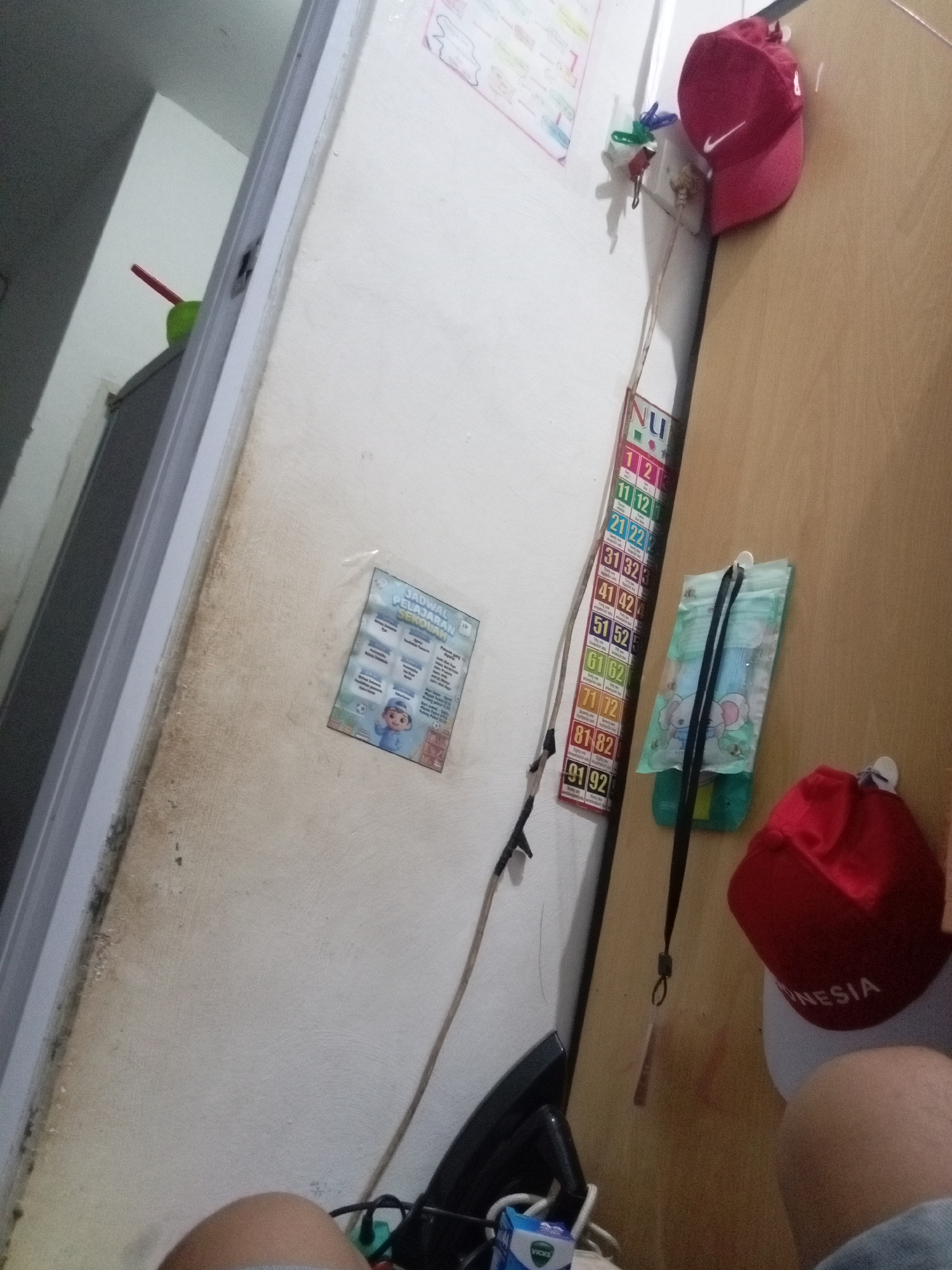 The image appears to be taken indoors, possibly in a residential setting, in Batam, Indonesia. The lighting suggests it is daytime, but the exact time is not discernible. The scene is cluttered, with various items affixed to or leaning against a light-colored, textured wall.
Dominating the right side of the frame is a light brown wooden door or cabinet. Hanging from it are two red baseball caps, one perched at the top and the other towards the bottom. The top cap has a white Nike logo. Below the top cap, a green pouch with an elephant print is attached to the door with a white adhesive hook and a black lanyard.
On the wall to the left, several items are visible. A vertically oriented chart or poster with numbered rows (e.g., 1-2, 11-12, 21-22, etc.) is attached. A small, laminated paper displaying a school schedule, with a cartoon illustration of a boy in traditional Indonesian attire, is also on the wall. A red pen or marker is inserted into a green object, possibly a holder, to the left of the schedule. A thin wire or cord runs diagonally across the wall.
In the lower left corner, a portion of a person's leg is visible, suggesting the photographer is present. A black electronic device, potentially a monitor or speaker, and a small blue and white box, possibly medication, are visible on a surface in the lower foreground. The wall itself shows signs of wear and discoloration, particularly along the left edge, with some dirt or mold present. The overall impression is of a functional, lived-in space with personal items displayed.