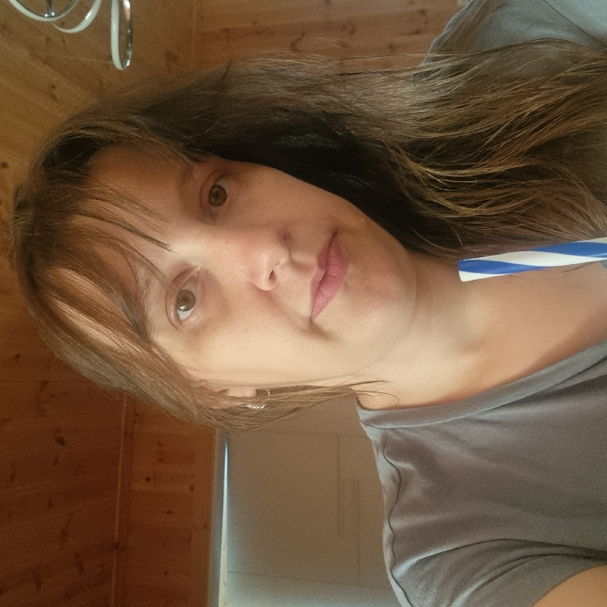 This is a close-up, rotated image of a person's face and upper body. The subject is a woman with long, brown hair and light-colored eyes. She is wearing a grey t-shirt. Her face is turned towards the camera, and she is looking slightly off-center. Her lips are parted, and she appears to be holding a blue and white striped object near her face. The background consists of wooden paneling and a lighter-colored wall. There are no visible text elements in the image. The lighting suggests it could be daytime indoors. The overall impression is that of a candid shot, possibly taken in a home setting.