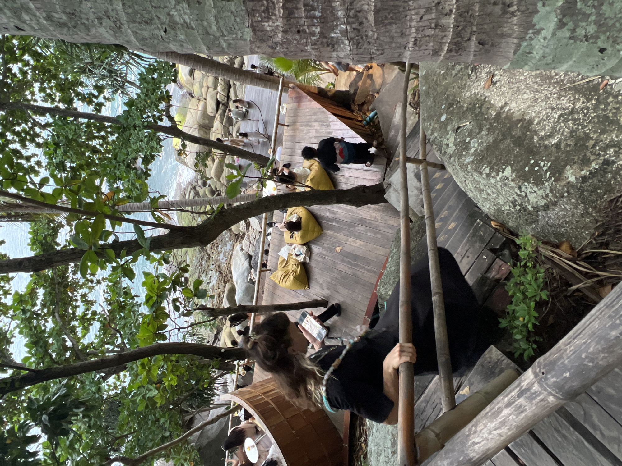 In Karon, Thailand, an outdoor restaurant area is depicted. A woman in a black dress is descending wooden stairs, holding a phone and looking down at it. She is leaning on a bamboo railing. Behind her, on a wooden deck, several people are seated on large yellow beanbag chairs. Some are looking at their phones, while others appear to be conversing. In the background, the ocean is visible, with waves crashing onto a rocky shore. The scene is surrounded by lush greenery and palm trees.