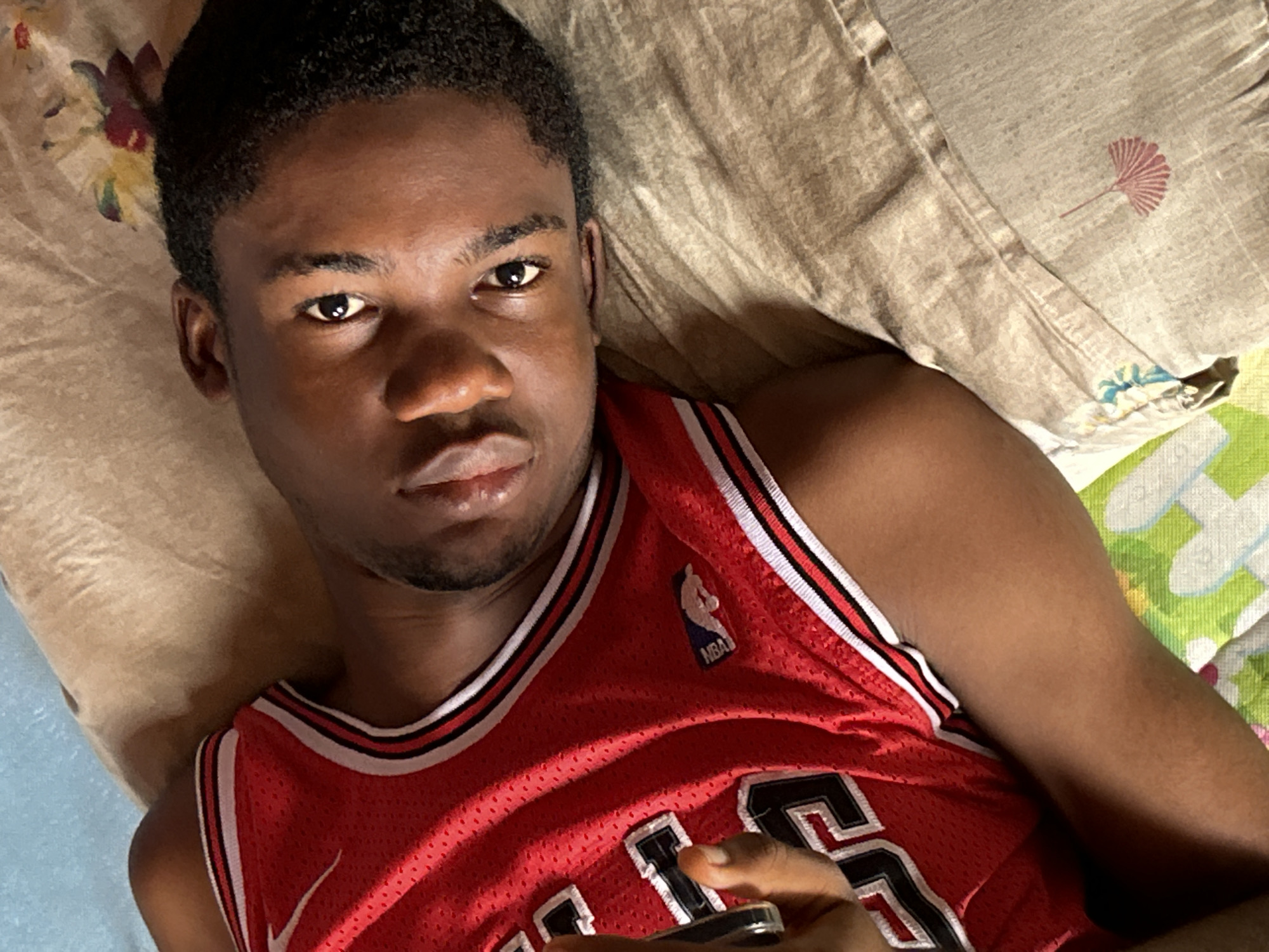 A young Black man is lying on his back in what appears to be a bed. He is wearing a red Chicago Bulls basketball jersey. The bed has a brown pillow and a multi-colored floral patterned sheet. The environment is not clearly identifiable as Ihiagwa, Nigeria, without additional context or identifiers in the image. He is holding a small object in his right hand, possibly a remote control. His expression is neutral, and he is looking at the camera.