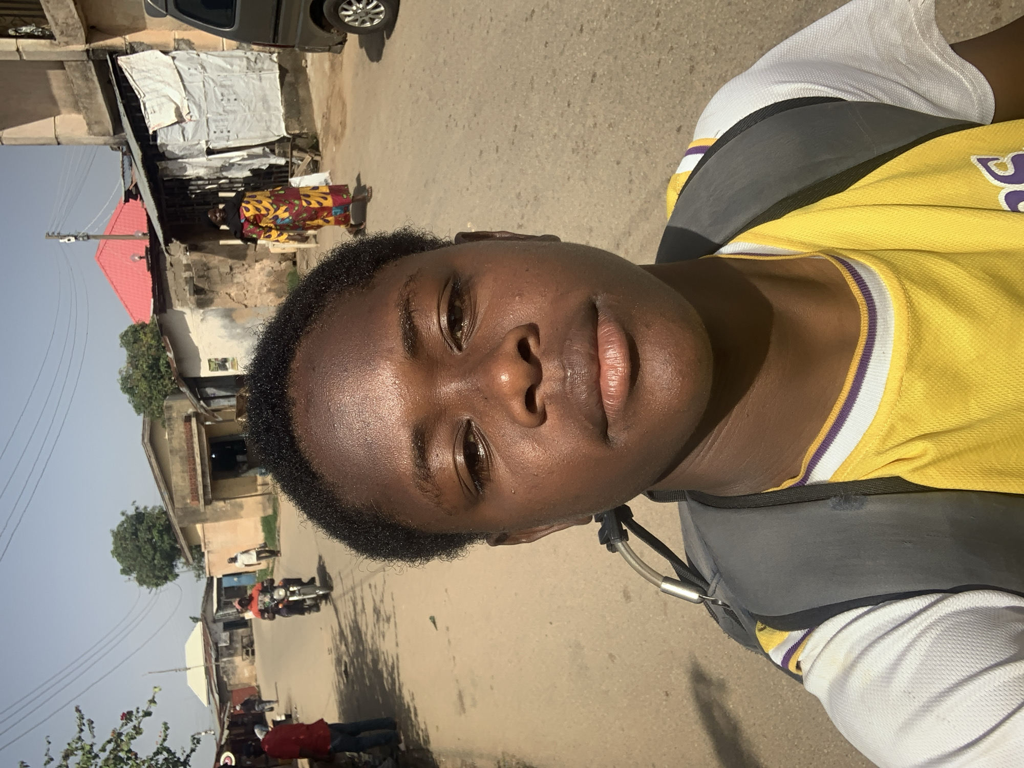 A close-up, ground-level shot faces directly toward a dark-skinned individual wearing a yellow and white collared shirt with purple trim and a grey backpack. The person is on a street in a busy city, likely Jos, Nigeria. The street is asphalt with light-colored dirt and appears worn. Behind the individual, the street leads away into the background, showing buildings of varying construction with people walking and riding motorcycles. There's also a parked vehicle on the upper left. The sky is visible in the upper left and is a light blue.