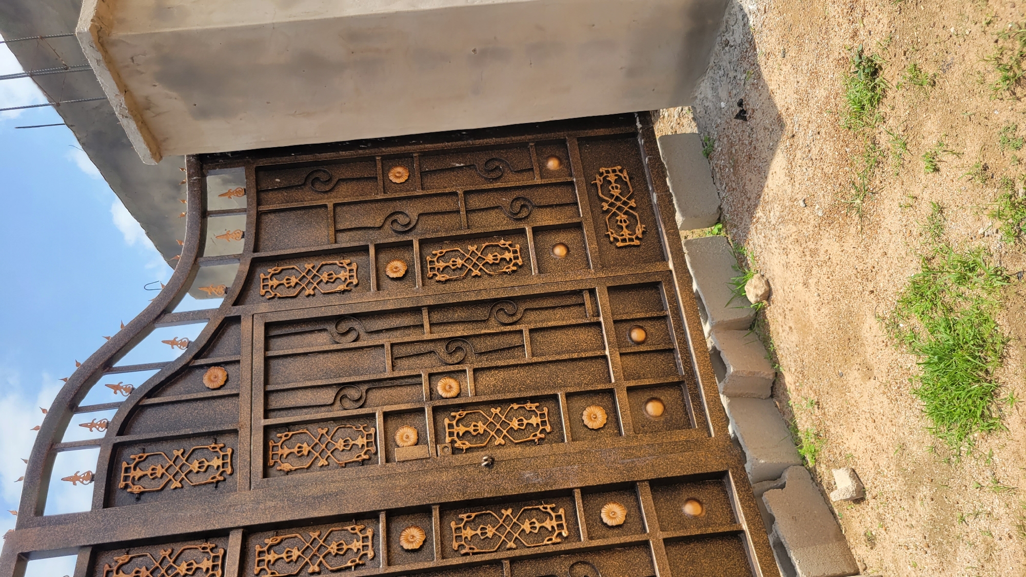 The image shows a closed, ornate brown metal gate with decorative gold patterns. The gate is partially open and leans to the right, revealing a glimpse of a construction site behind it. The construction site appears to have a concrete pillar on the right side of the gate, and a gray wall or structure with rebar sticking out of it in the background. The ground in front of the gate is covered with sand, small pebbles, and patches of green grass. Several concrete blocks are placed along the bottom of the gate, likely as a temporary foundation or support. The sky above is blue with scattered white clouds. The scene is consistent with a construction or renovation project in Mayo Dasa, Nigeria, given the visible architectural elements and the overall environment.